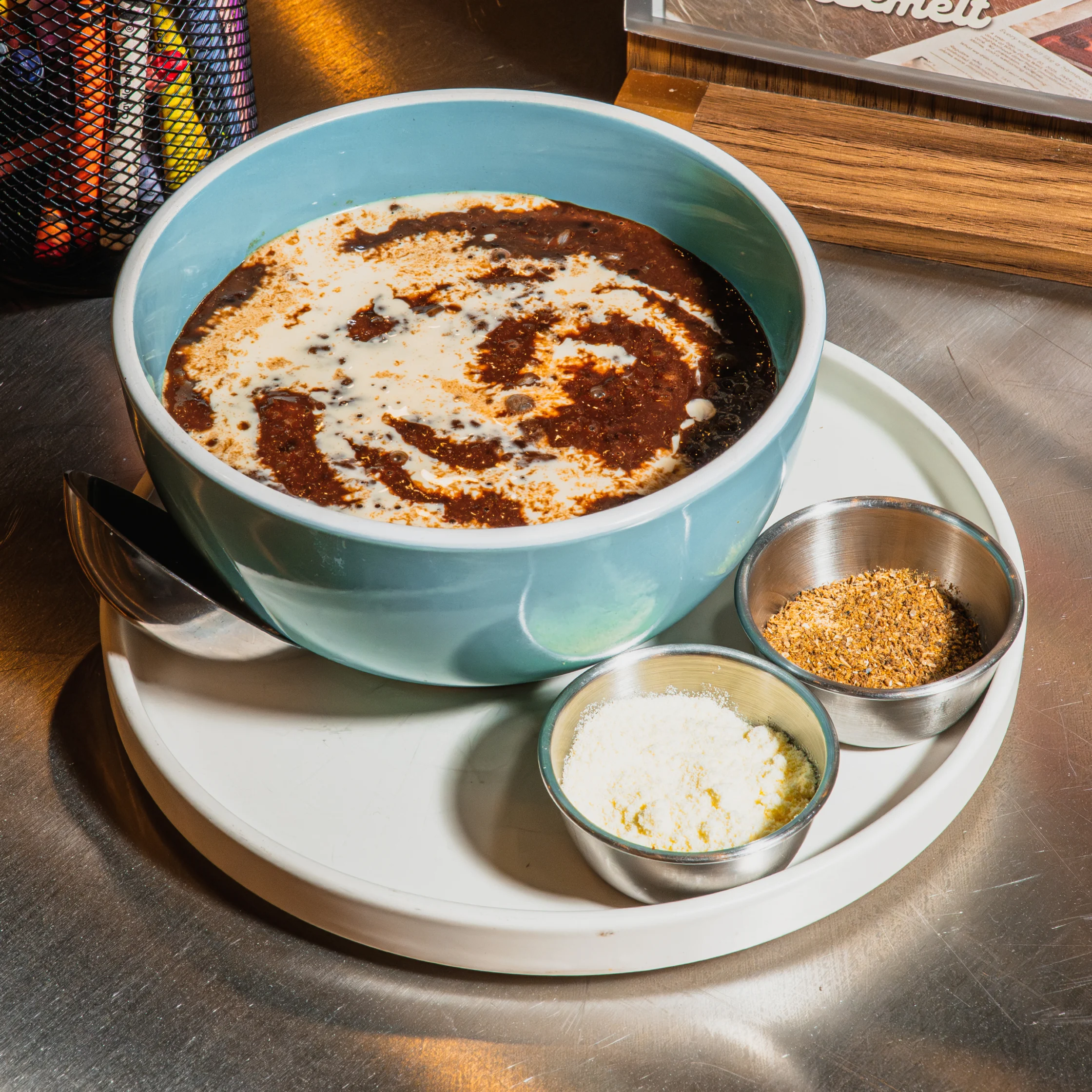 A blue bowl of creamy rice porridge topped with dark toppings sits on a white tray beside small cups of seasoning, ready to enjoy in a cozy café.