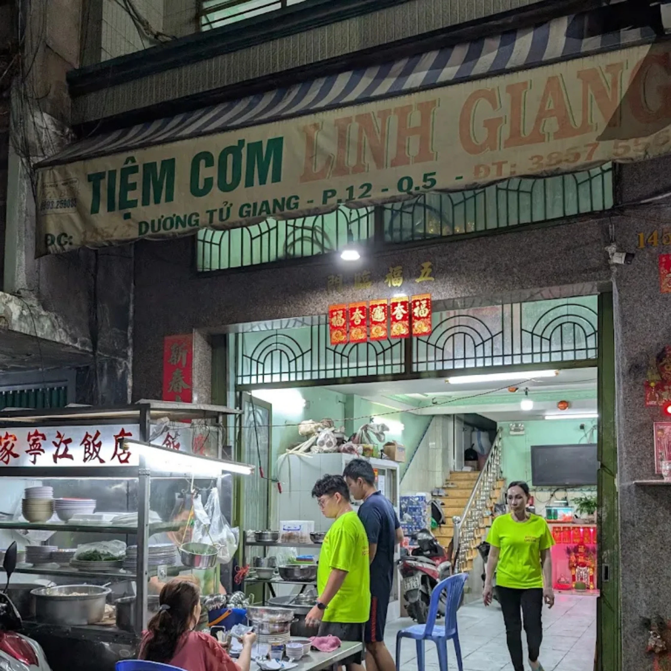 Nighttime Vietnamese street-side eatery, with diners at plastic tables and staff in bright green shirts beneath a weathered “Tiệm Cơm Minh Giang” sign.