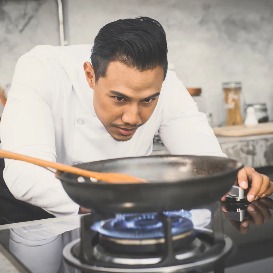 A focused chef in a white jacket adjusts a gas burner beneath a frying pan in a professional kitchen, preparing to cook.