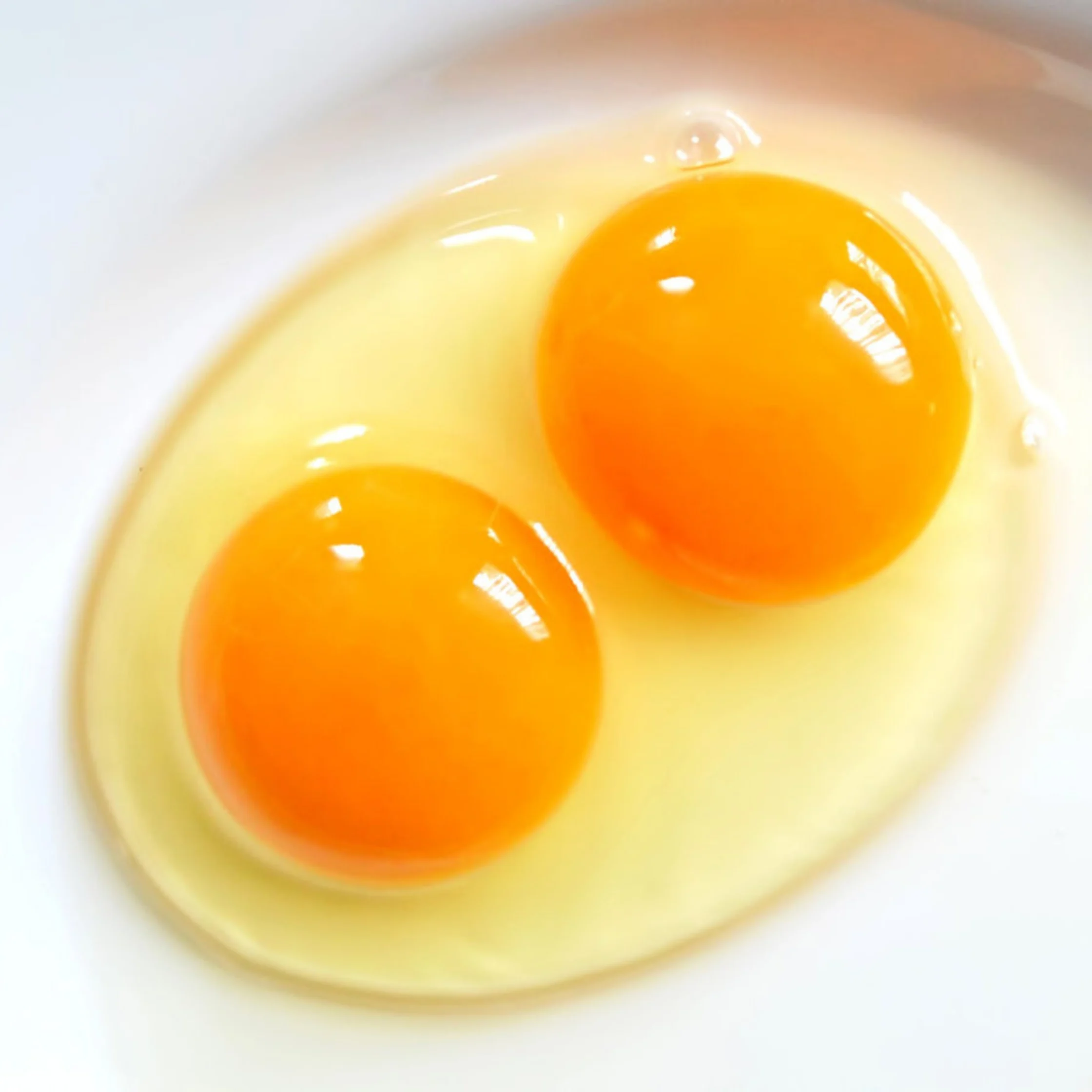 Two bright orange egg yolks sit in translucent egg white on a white plate, creating a fresh, simple close-up with a clean, appetizing feel.