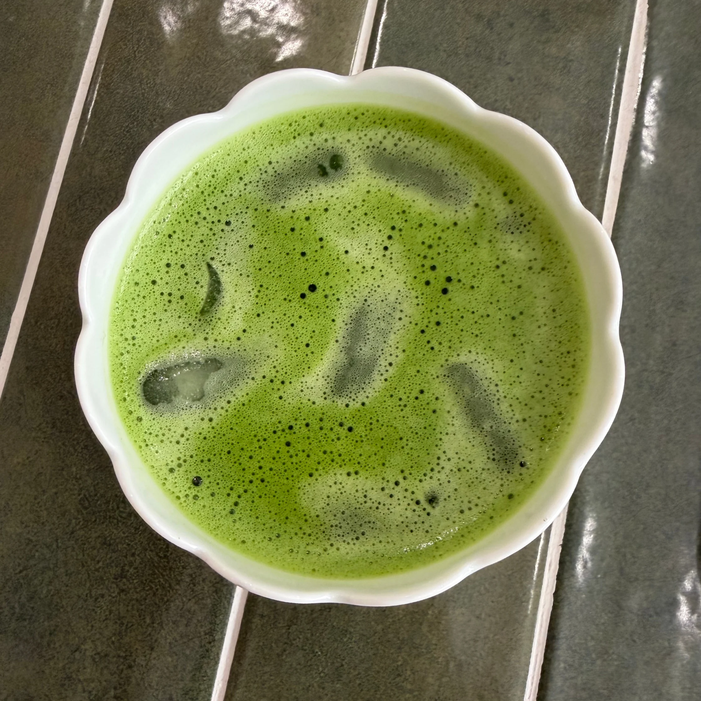 A white scalloped bowl holds bright green, frothy matcha tea with tiny bubbles on a dark tiled surface, creating a calm, fresh mood.