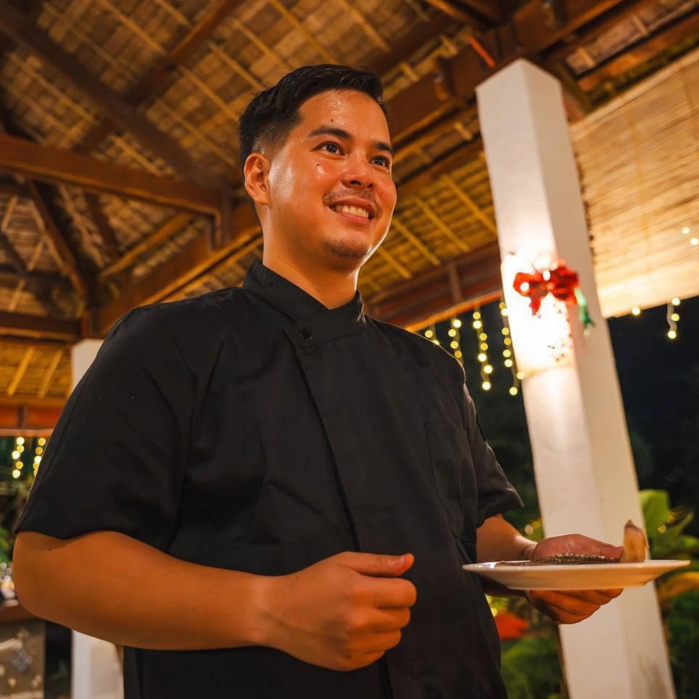 A smiling man in a black shirt holds a plate beneath a warmly lit thatched roof, with festive string lights creating a welcoming evening atmosphere.