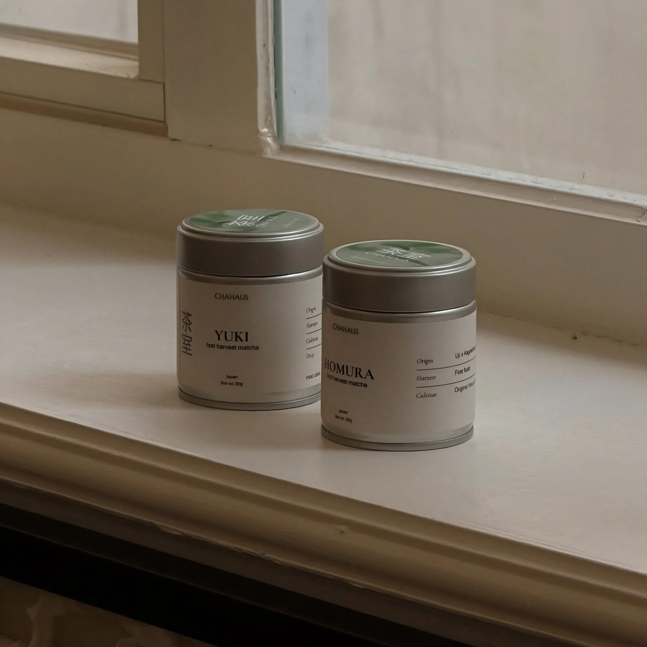 Two small gray tea tins labeled Yuki and Homura sit on a sunlit windowsill, their calm, minimalist packaging lending the quiet indoor scene a serene tone.
