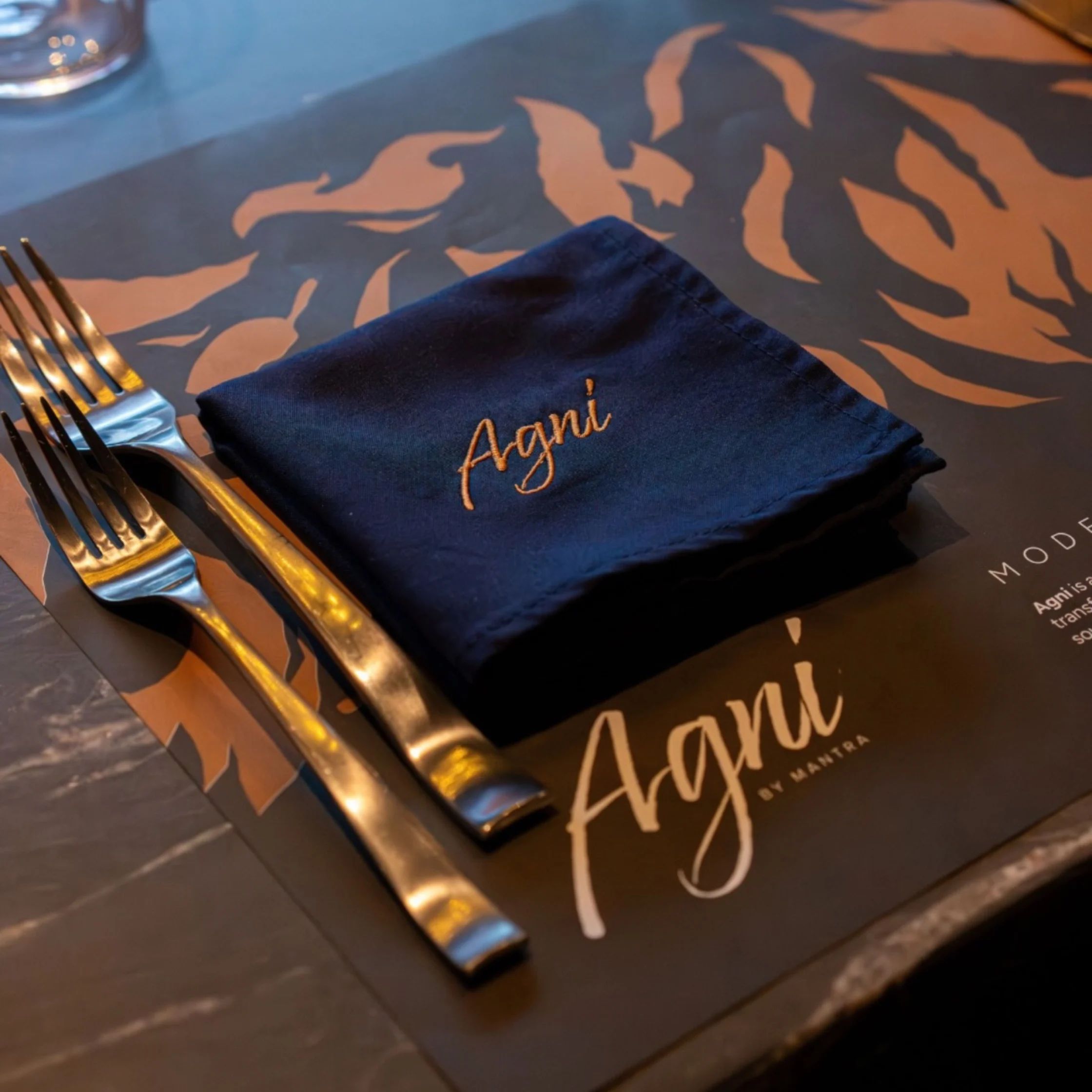 A stylish restaurant table setting with a navy napkin embroidered “Agni,” gold cutlery, printed menu, and water glasses, conveying an elegant, warm mood.