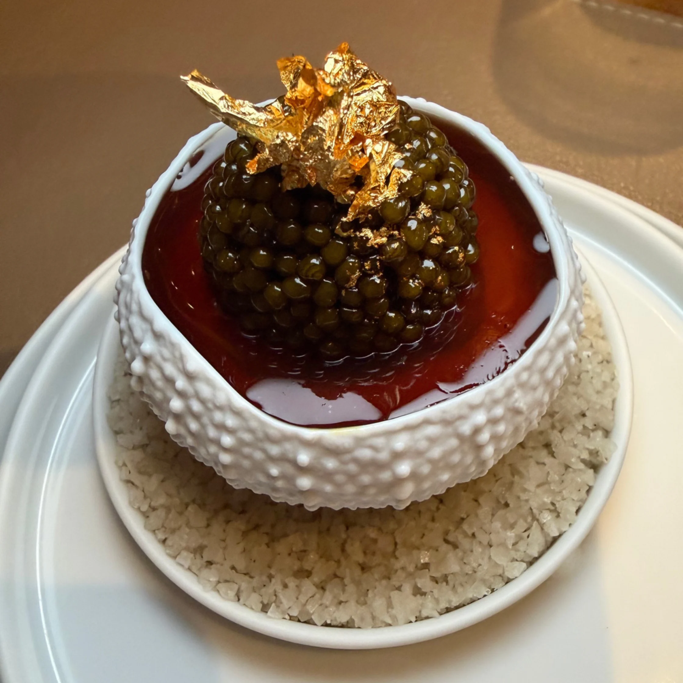 A dramatic gourmet dessert features glossy black caviar-like pearls and edible gold leaf in a white textured bowl, surrounded by red sauce and coarse sugar.