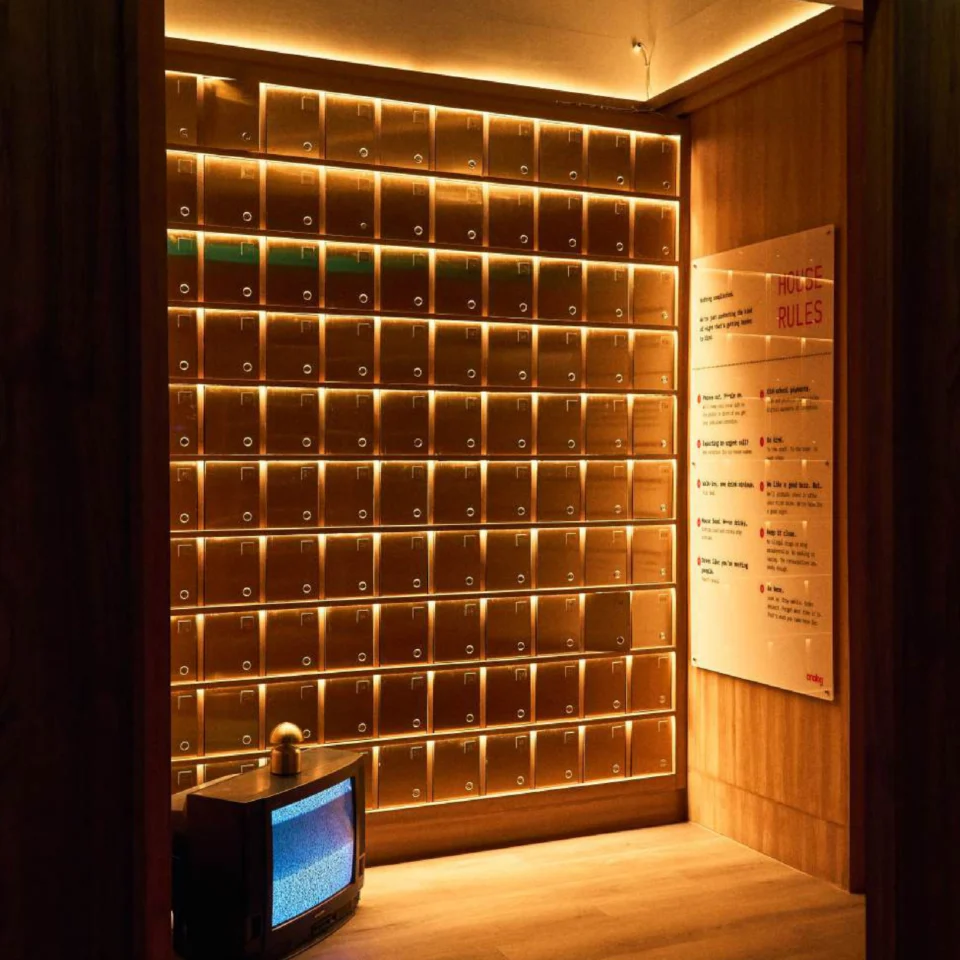 A warm, amber-lit room lined with rows of small metal lockers, a vintage CRT television in the corner, and a rules sign on the wall.