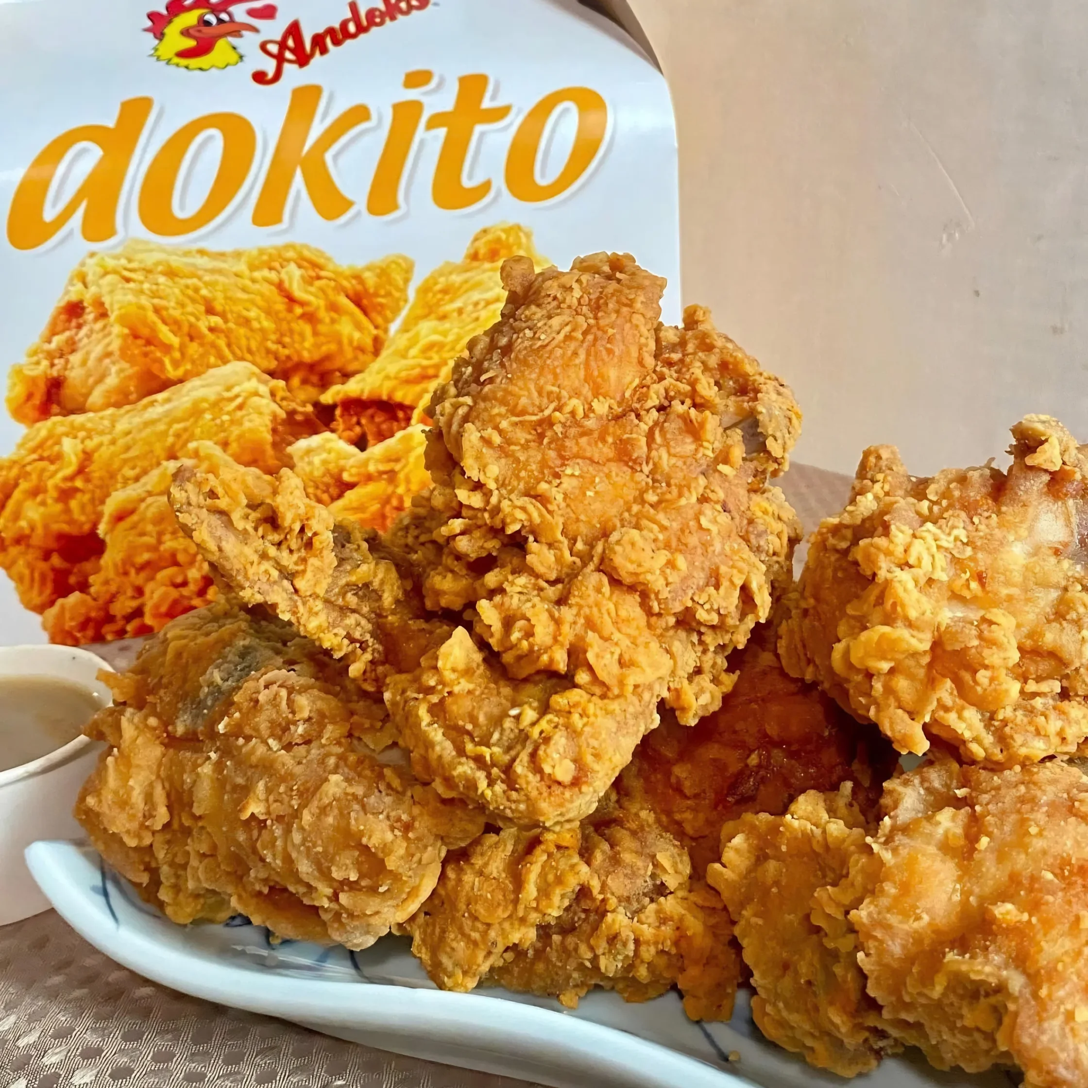 A platter piled with crispy, golden-brown fried chicken sits in front of a “Andok’s Dokito” package, conveying a hearty, appetizing meal.