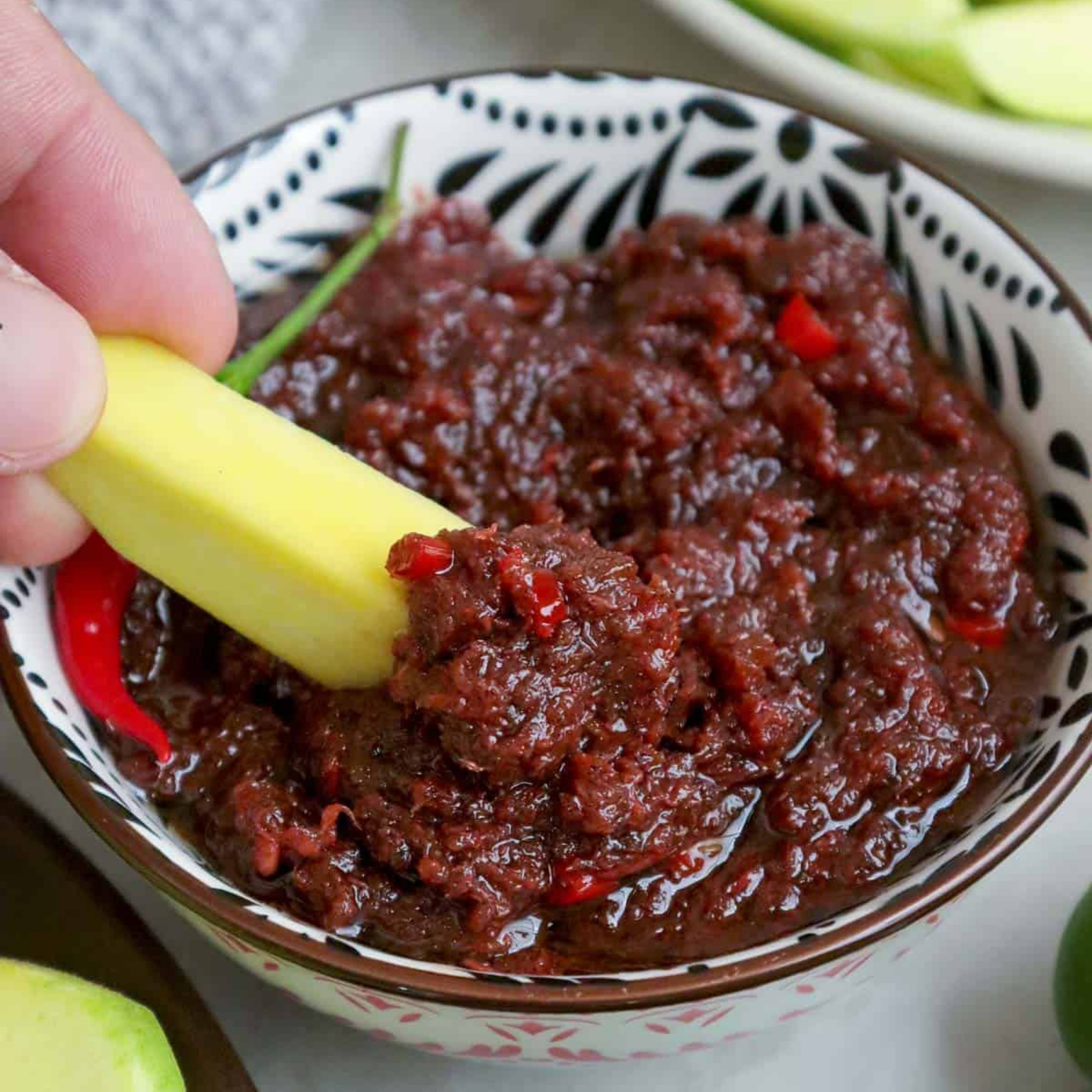A hand dips a yellow vegetable stick into chunky red-brown bagoong in a patterned bowl, with red pepper pieces and fresh vegetables nearby; appetizing, casual snack.