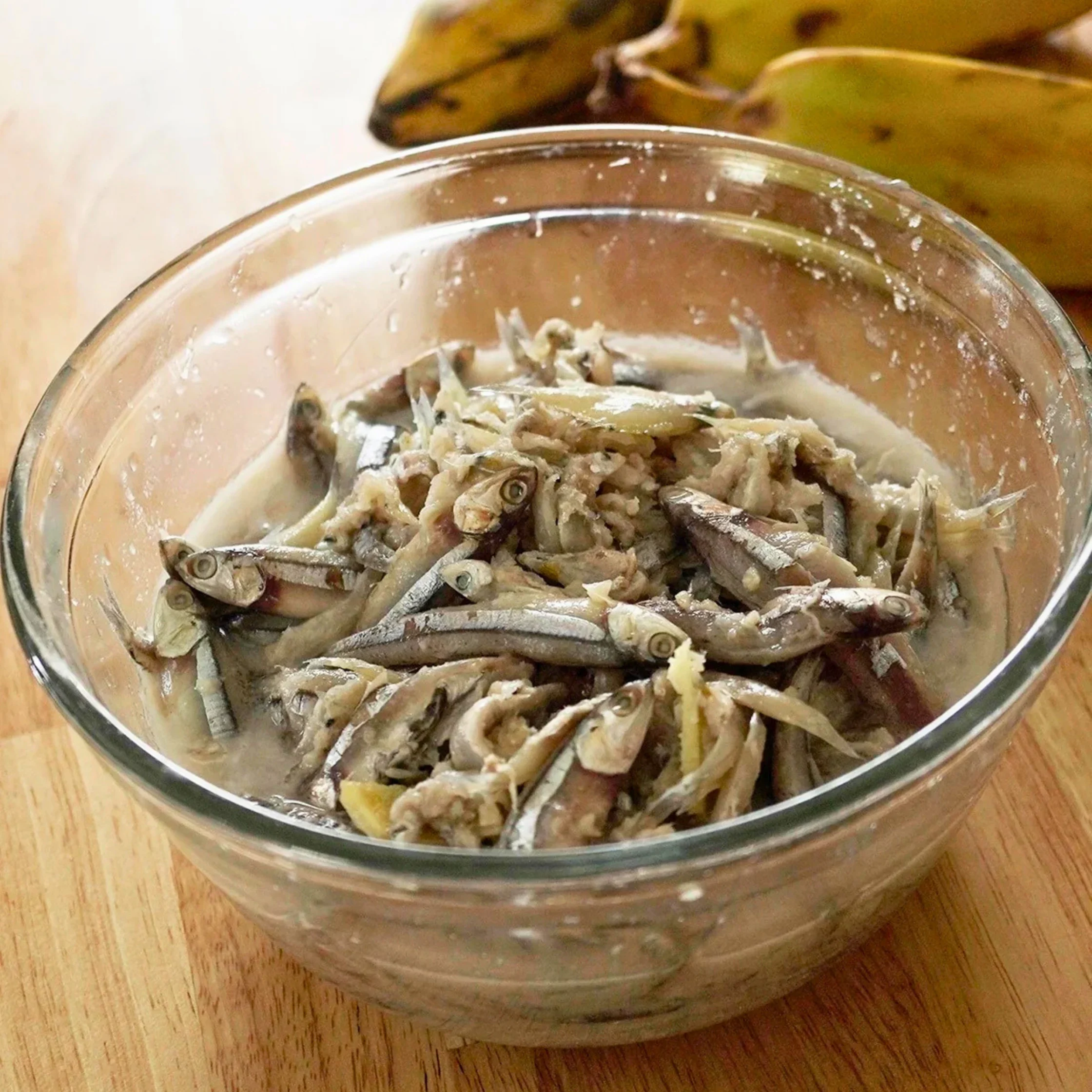A glass bowl holds small whole fish mixed with a pale, creamy sauce on a wooden counter, with ripe bananas behind it; the rustic dish looks savory.