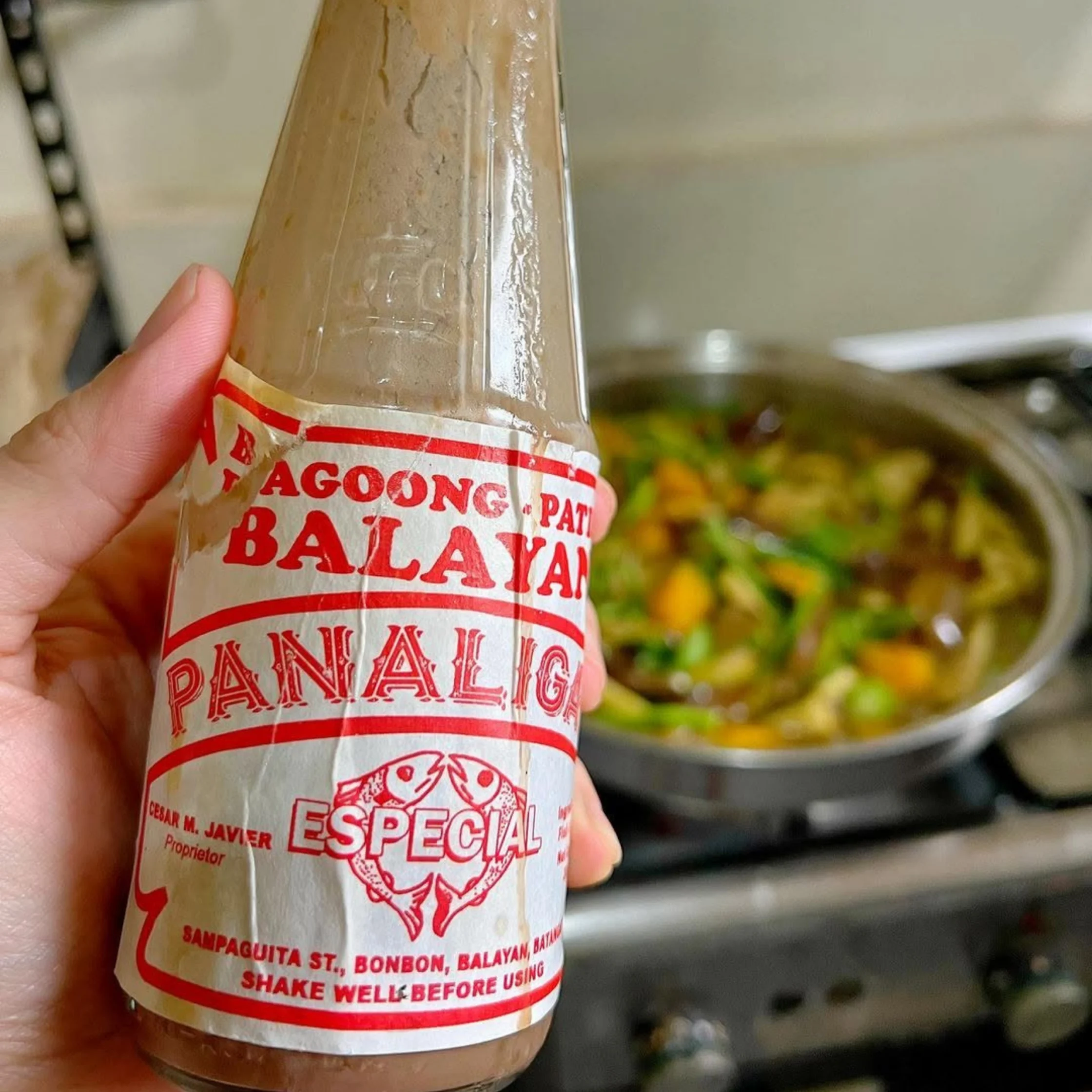 A hand holds a glass bottle of red-labeled Panalang special sauce beside a pan of simmering vegetables, evoking a warm Filipino home-cooking scene.