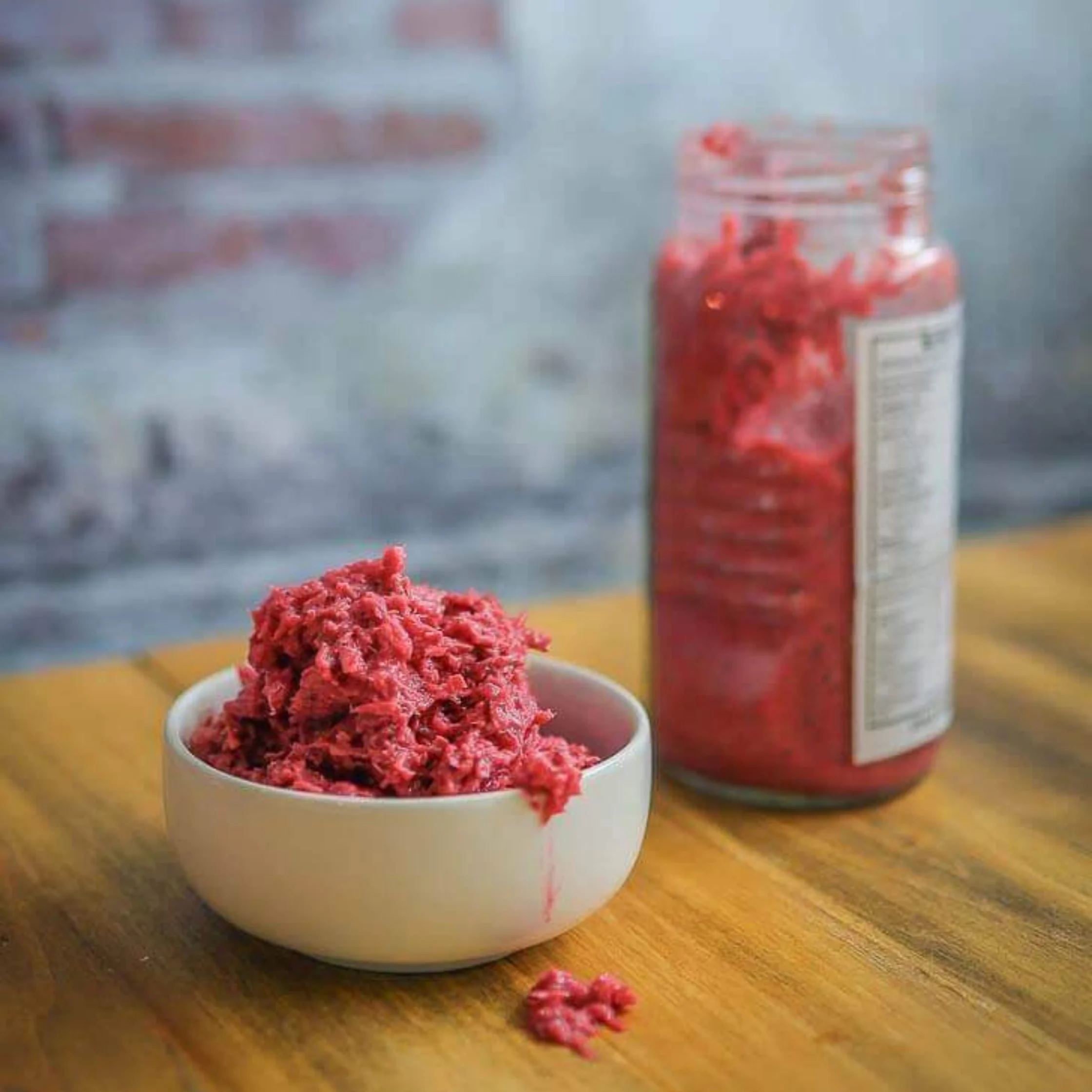 A bowl of vivid red minced shrimp paste sits on a wooden counter beside an open glass jar of the same condiment, suggesting a bold, spicy kitchen staple.
