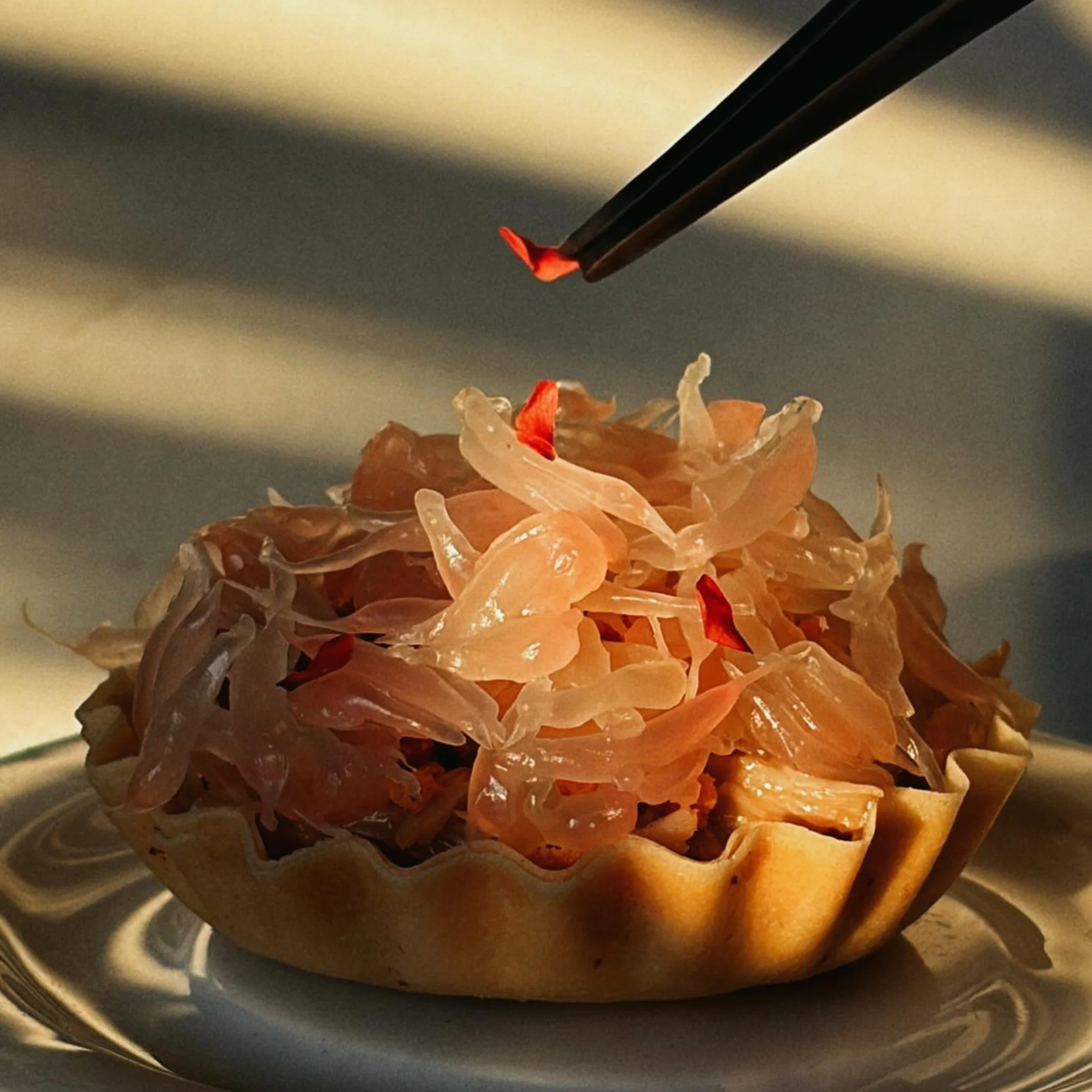 Warm sunlight highlights a golden tart piled with translucent orange shreds and red garnish, as black chopsticks lift a small piece; an elegant, appetizing food scene.