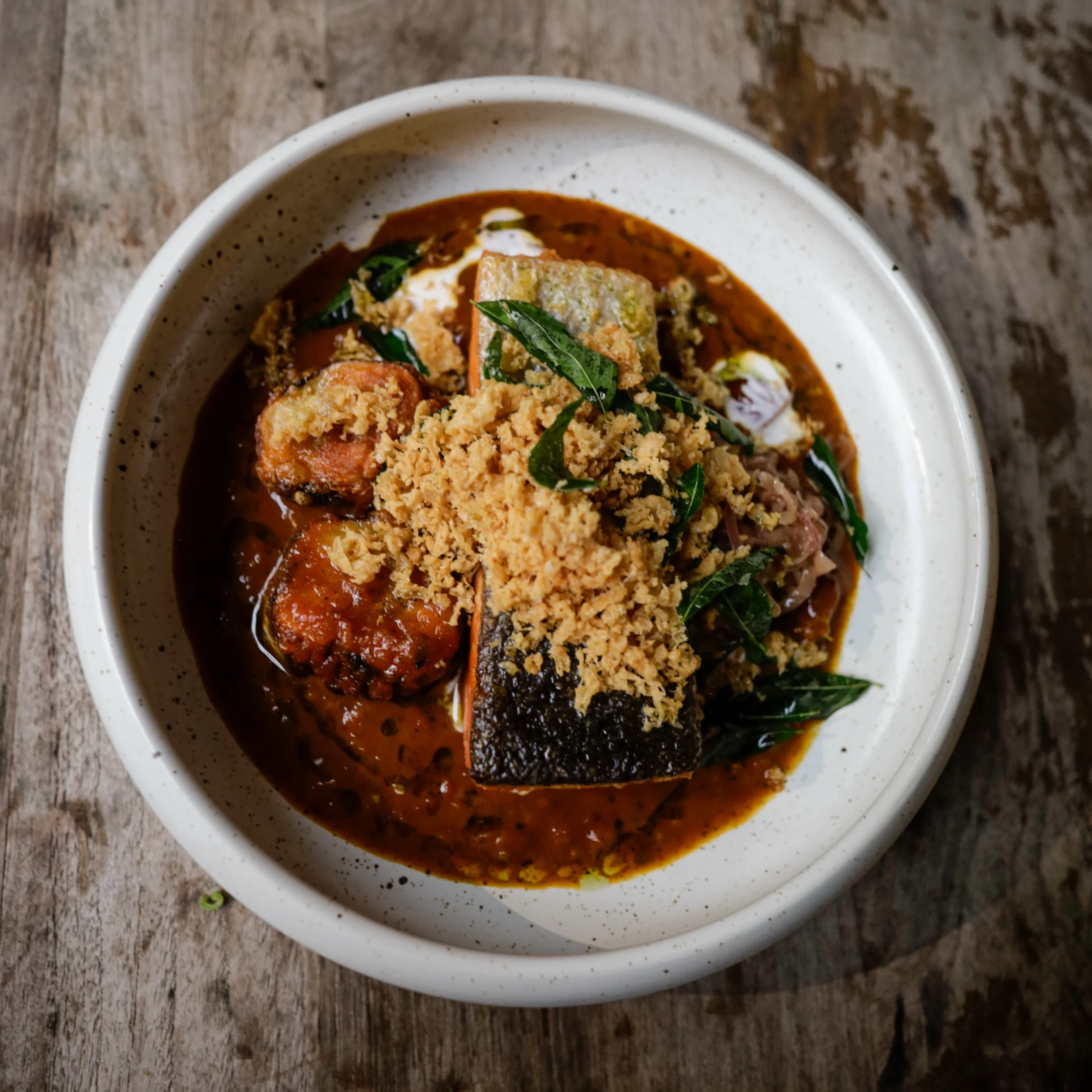 Overhead view of a white bowl filled with rich reddish-brown fish curry, topped with crispy golden crumbs and fresh green herbs, on a rustic wooden table.