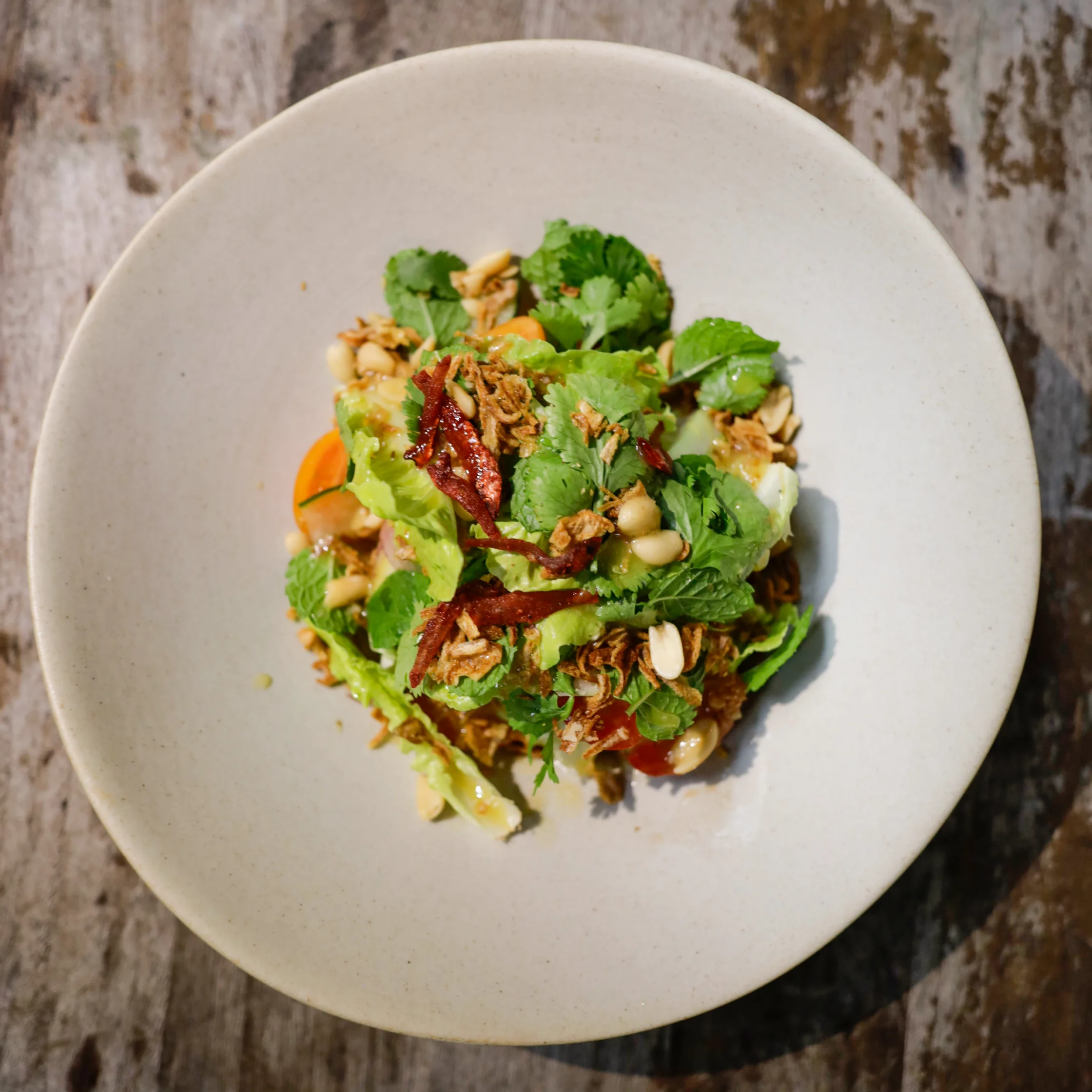 Overhead view of a vibrant salad on a cream ceramic plate, with leafy greens, chickpeas, herbs, peanuts, and crisp red chili. Fresh and inviting.