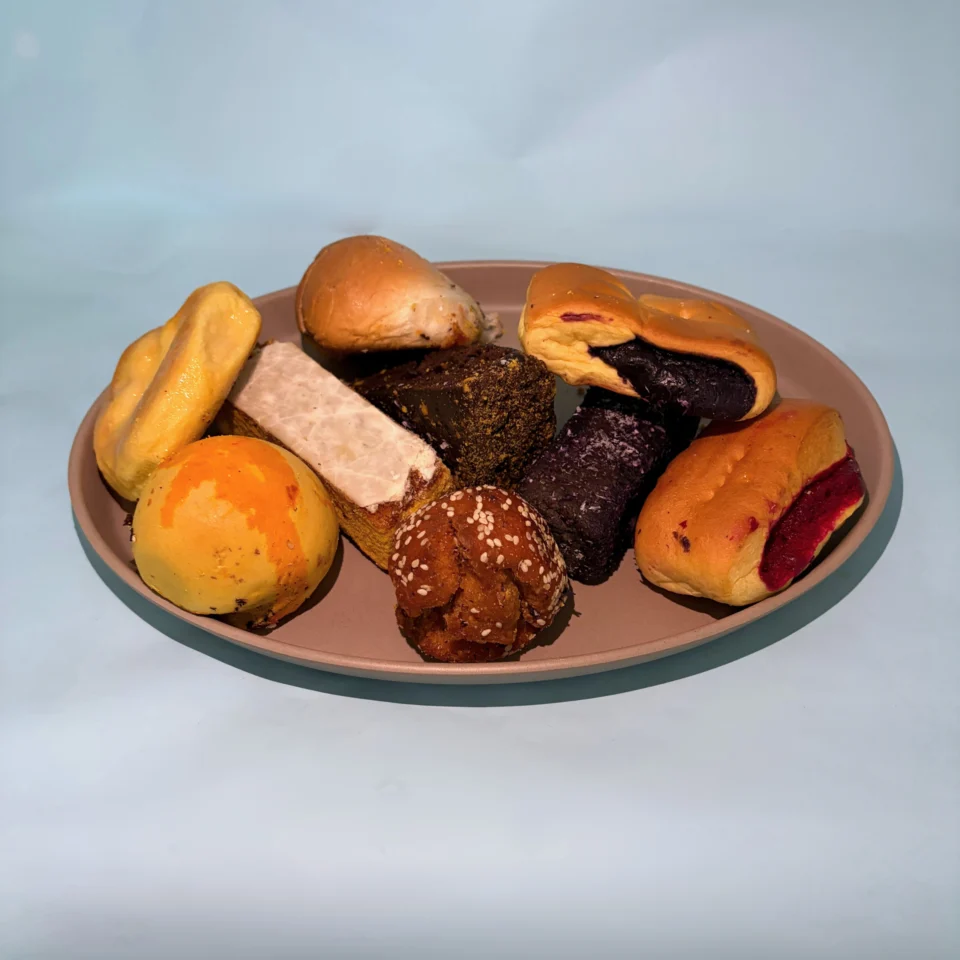 An oval plate holds assorted pastries, rolls, and dark and light cakes against a pale blue background, creating a warm, inviting still life.