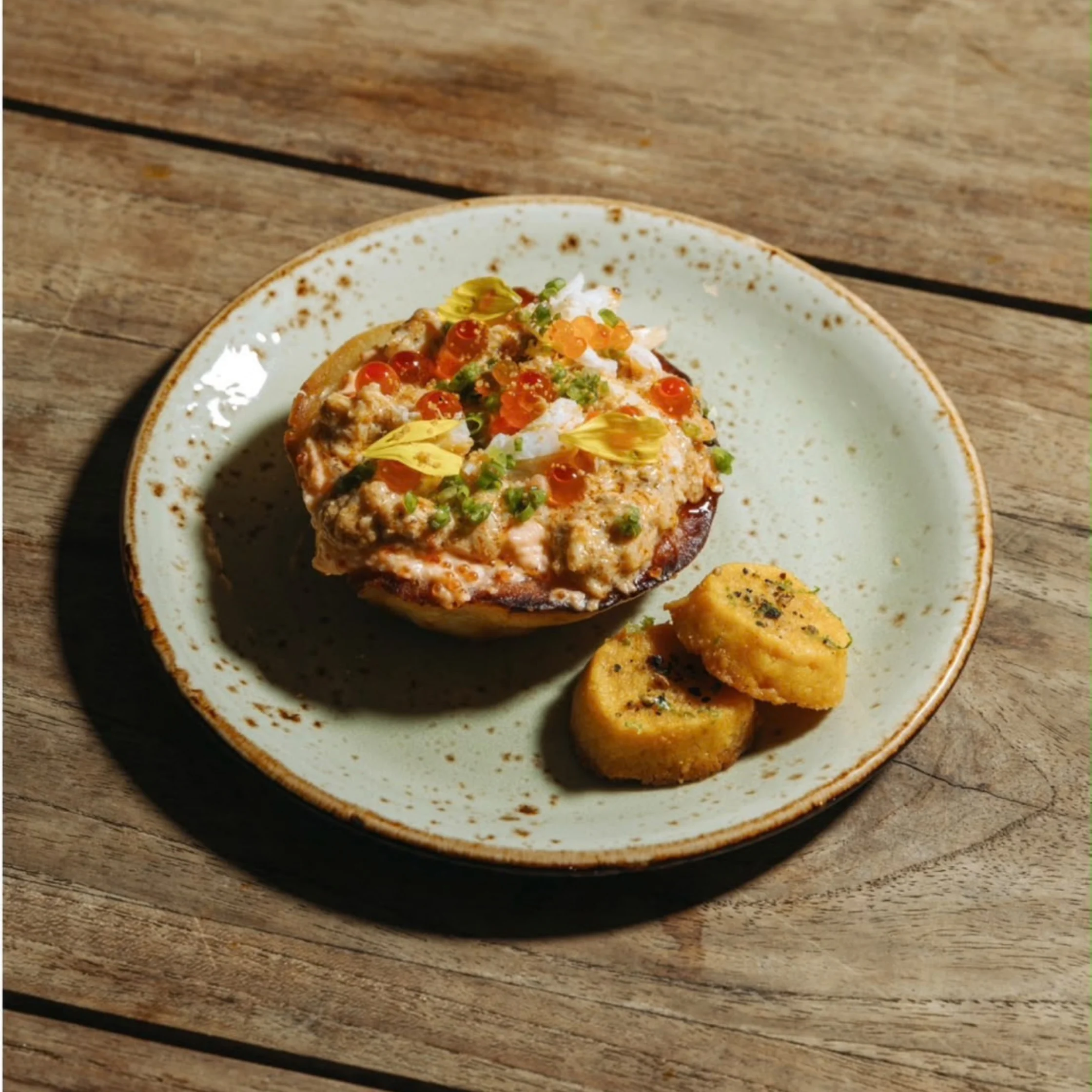 A savory stuffed eggplant topped with herbs, diced vegetables, and cheese sits on a speckled ceramic plate beside two golden rounds, in warm rustic sunlight.