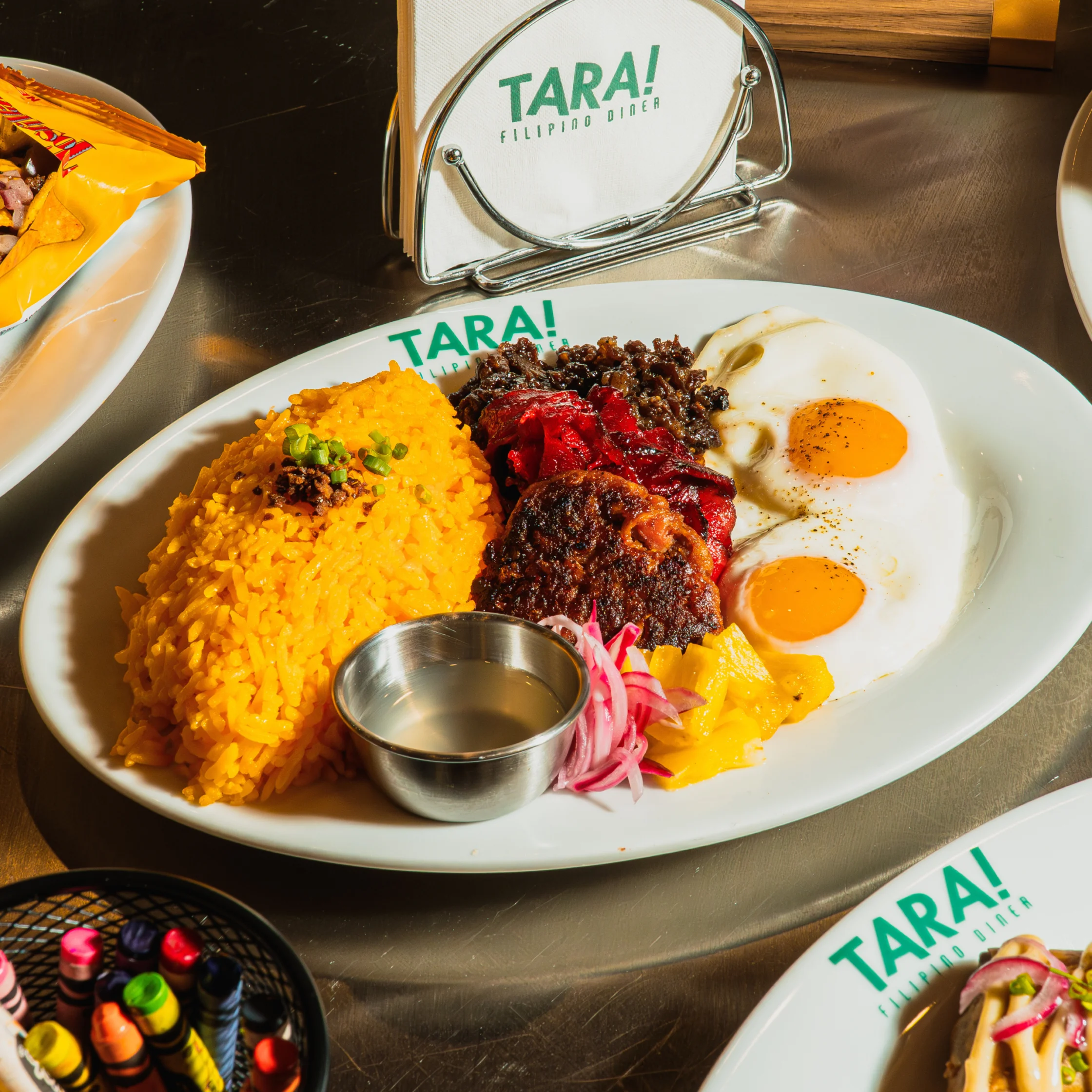 A Tara! Filipino diner platter with golden garlic rice, two fried eggs, longganisa, tocino, pickled onions, and dipping sauce. Warm, hearty meal.