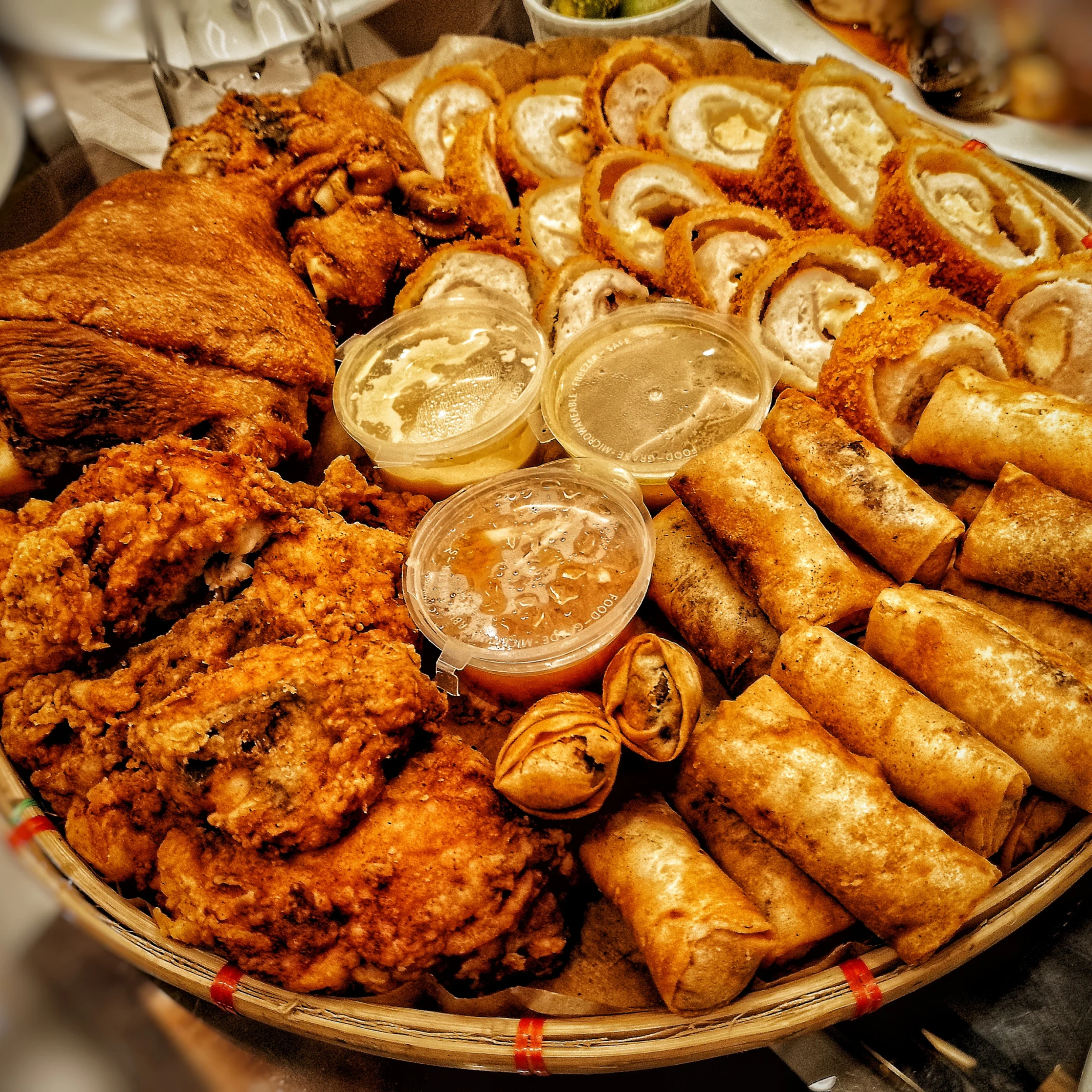 A platter piled with crispy fried chicken, golden spring rolls, sliced savory rolls, and three cups of dipping sauce, creating a hearty, festive spread.
