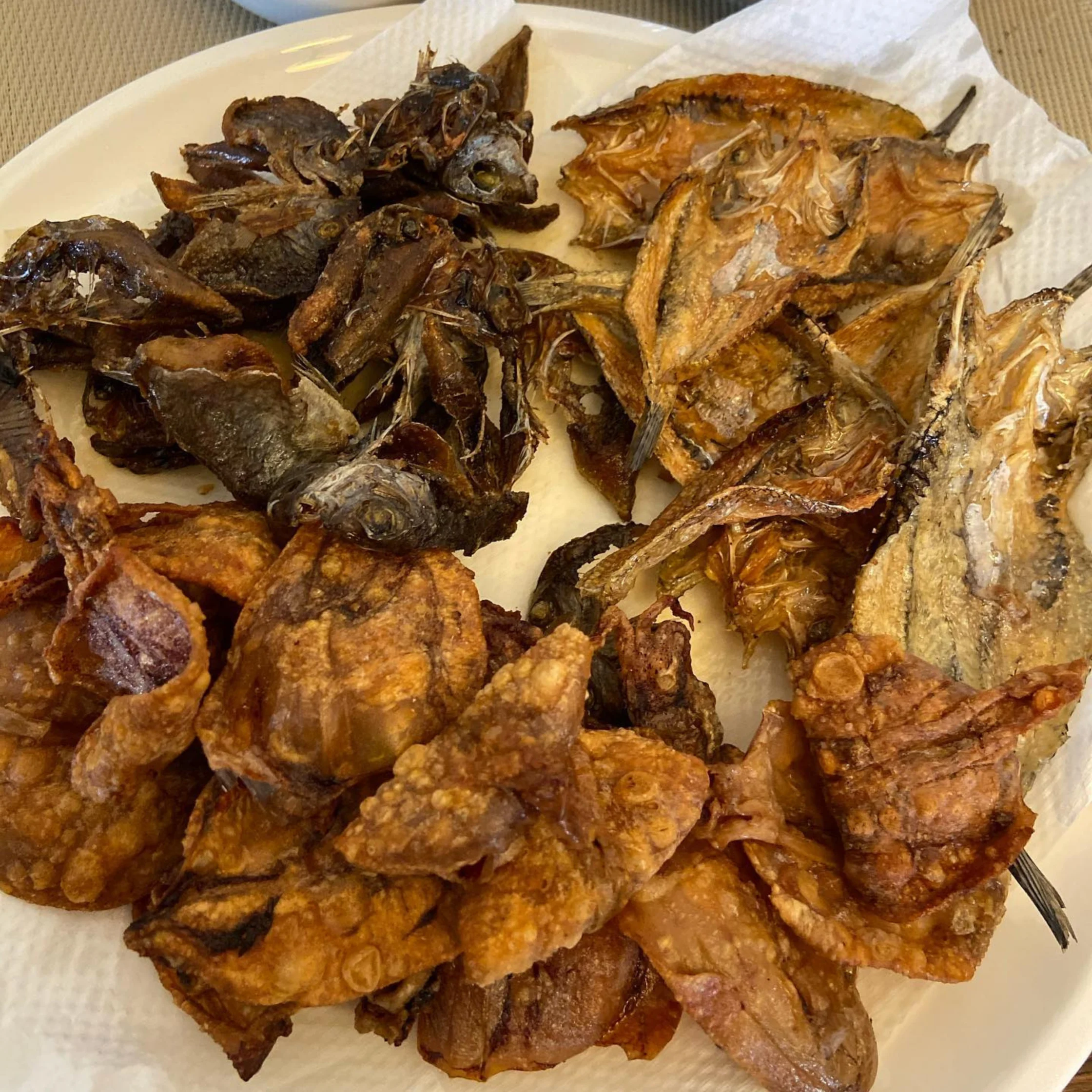 A white plate holds assorted fried and dried fish, from dark whole fish to golden crispy pieces, creating a hearty, rustic meal.