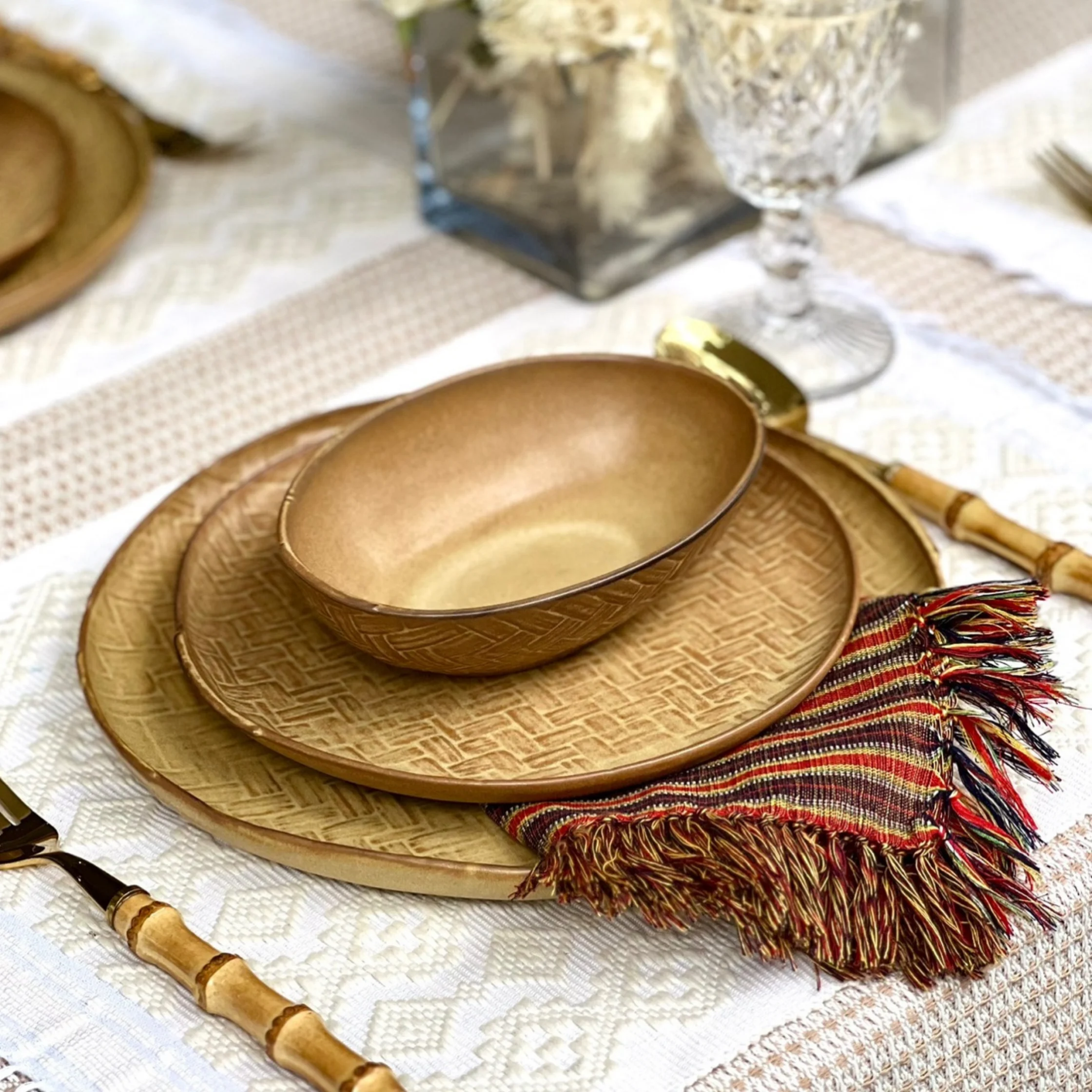 Gold-toned woven placemats and a matching bowl sit on a white patterned tablecloth beside a fringed red-and-gold napkin, creating an elegant, warm tablescape.