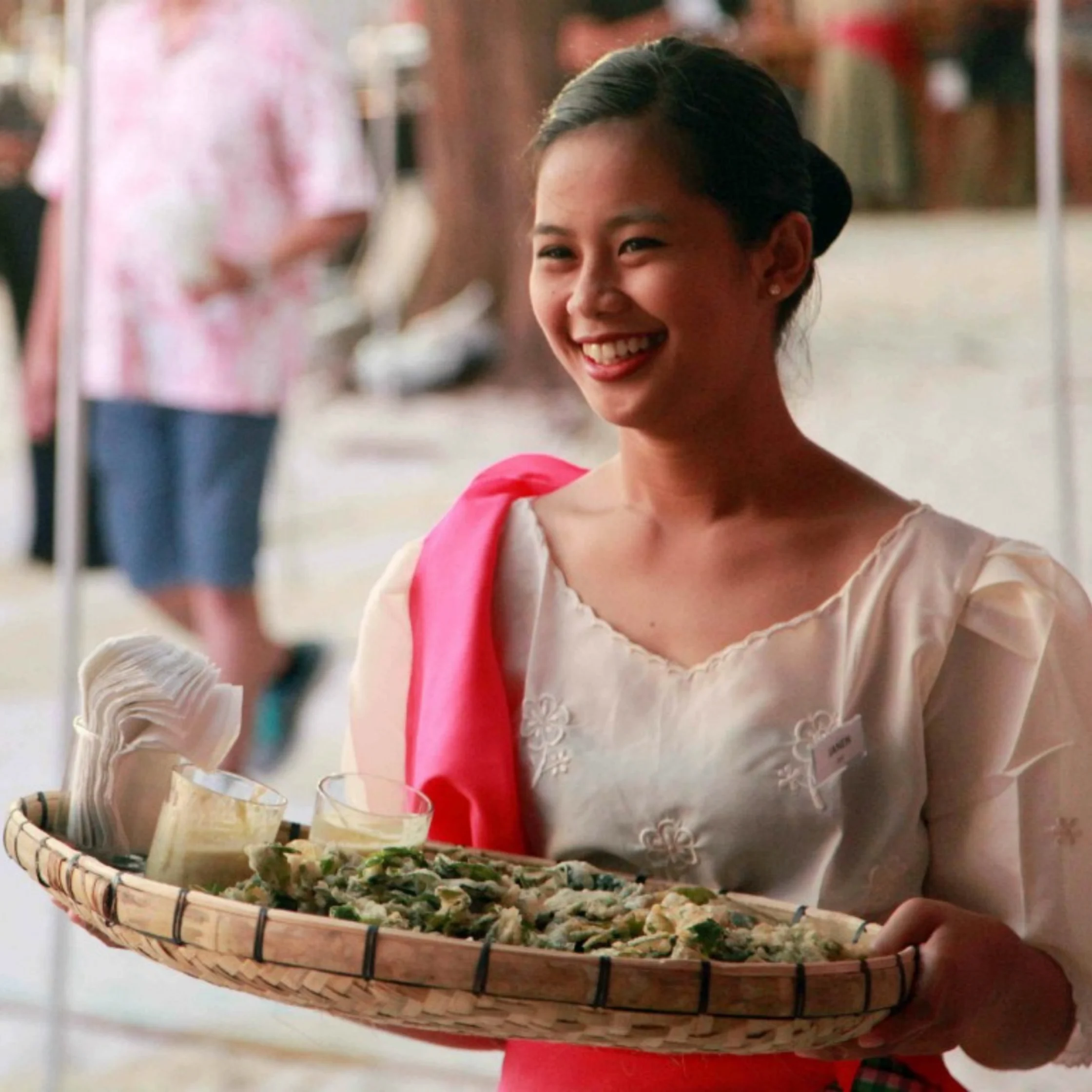 A smiling young woman in traditional white clothing and a bright pink sash holds a woven tray of offerings at an outdoor market, conveying warmth and welcome.