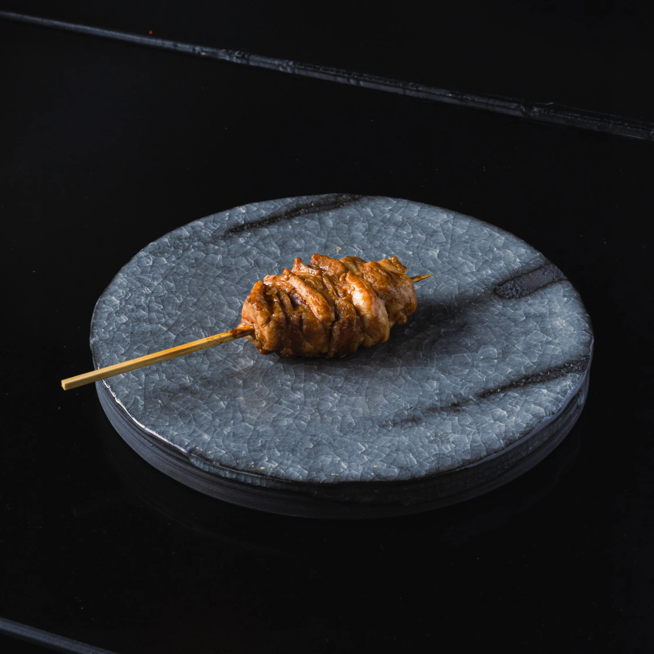 A single grilled meat skewer rests on a round gray stone plate against a dark background, creating a moody, minimalist food presentation.