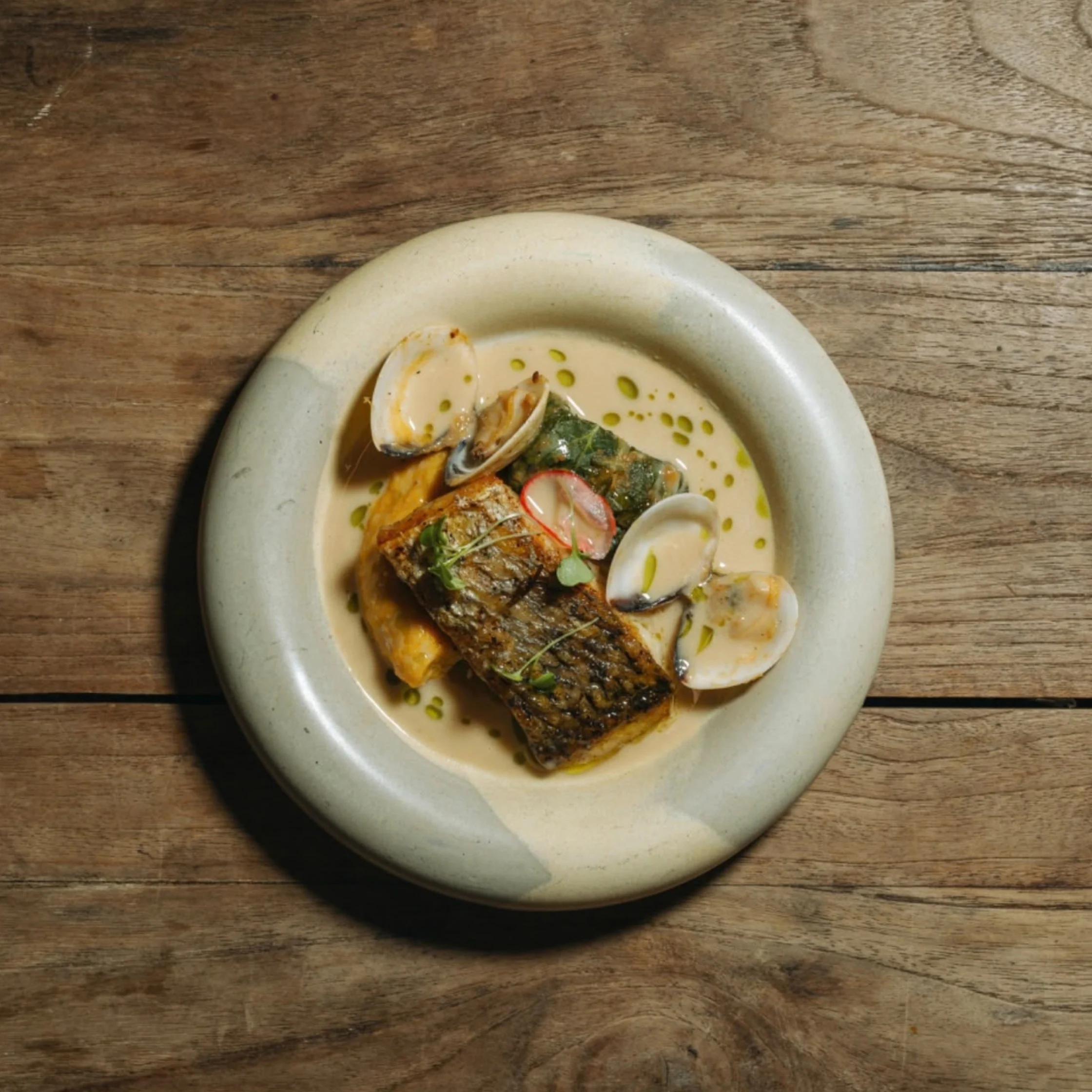 Overhead view of grilled fish fillet, clams, and vegetables in creamy sauce on a pale ceramic plate atop a rustic wooden table; warm, inviting mood.