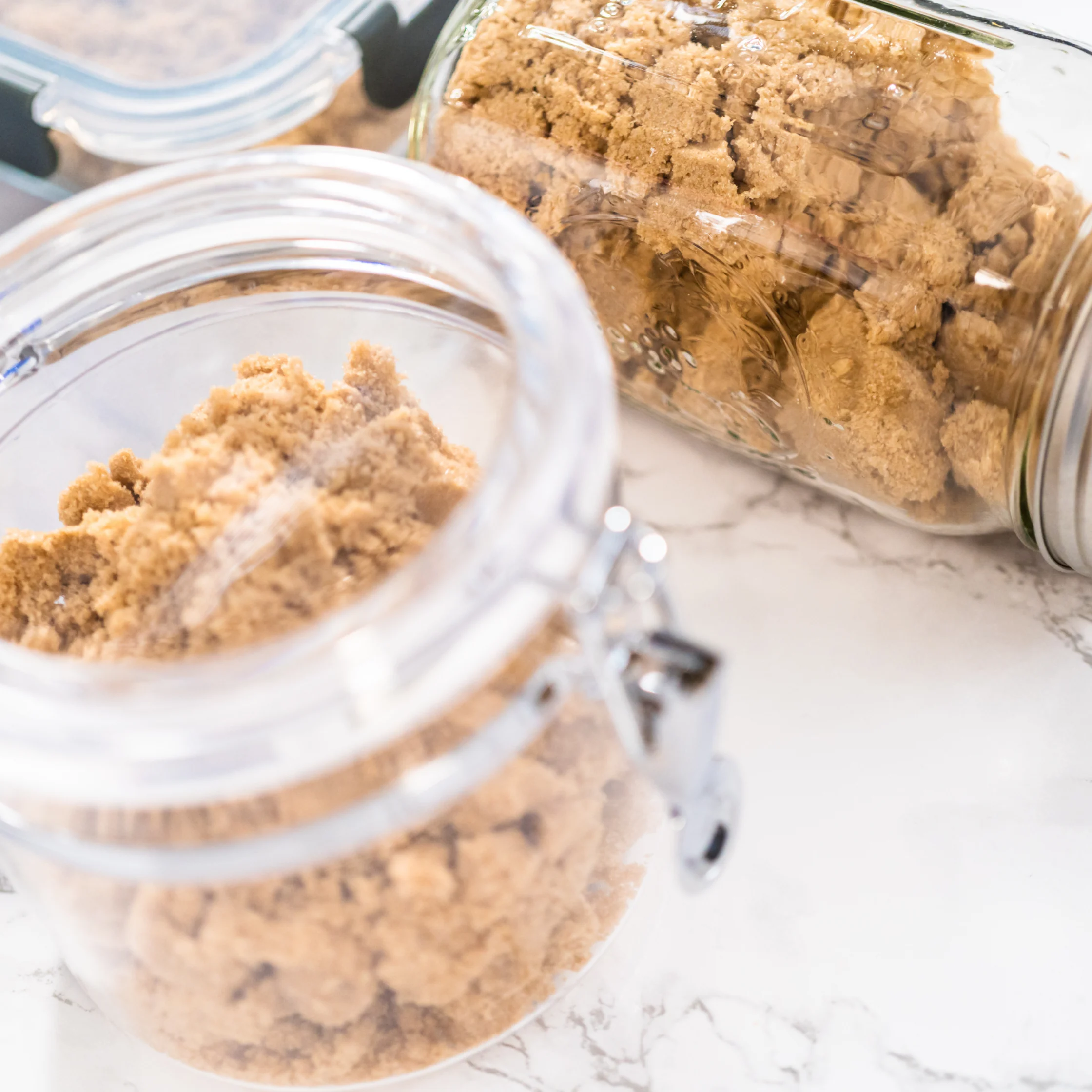 Two clear glass jars hold crumbled brown sugar on a bright marble counter, with a scoop in the open jar; a warm, tidy kitchen scene.