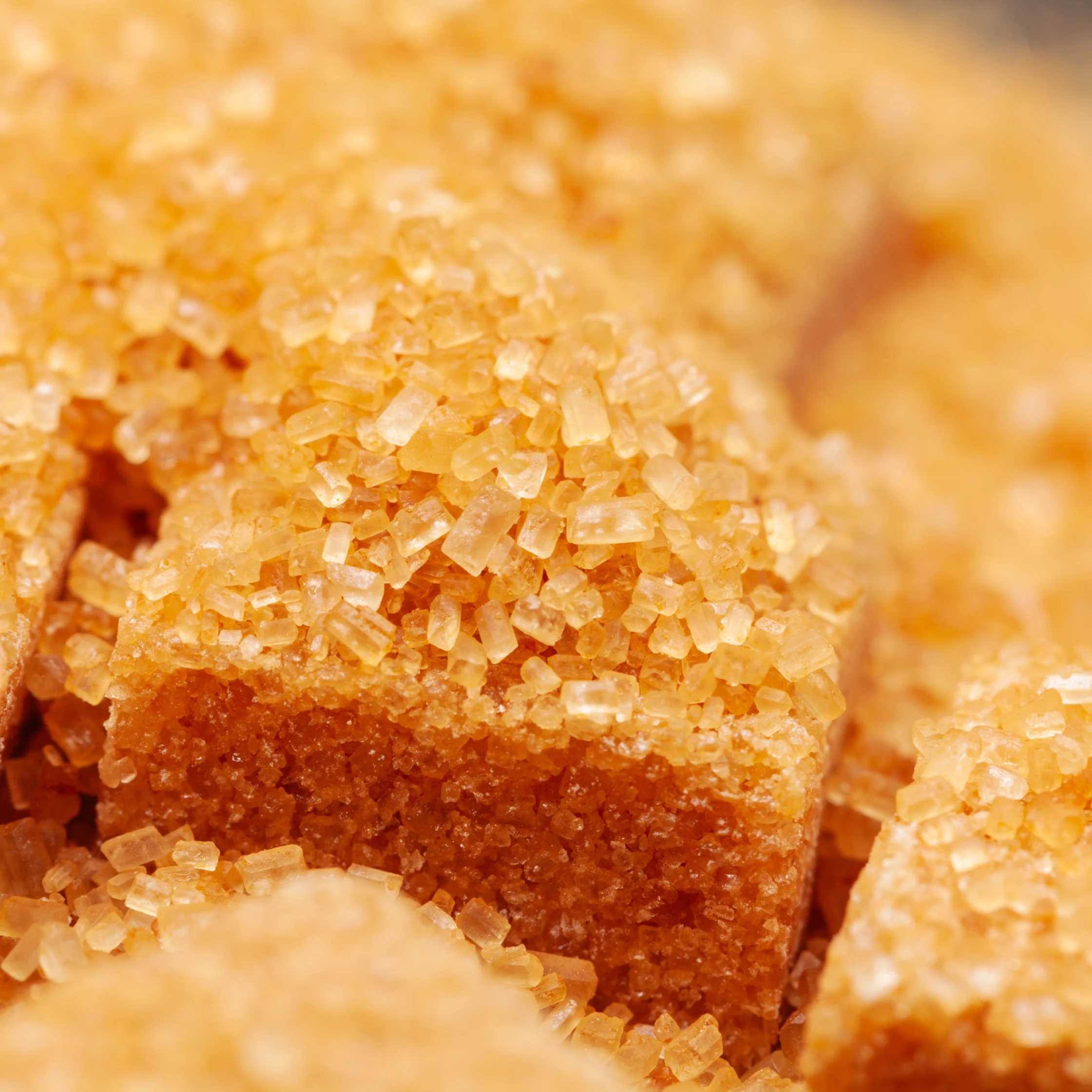 Macro close-up of golden-brown sugar cubes coated in sparkling coarse crystals, with more cubes softly blurred behind them, evoking warmth and sweetness.