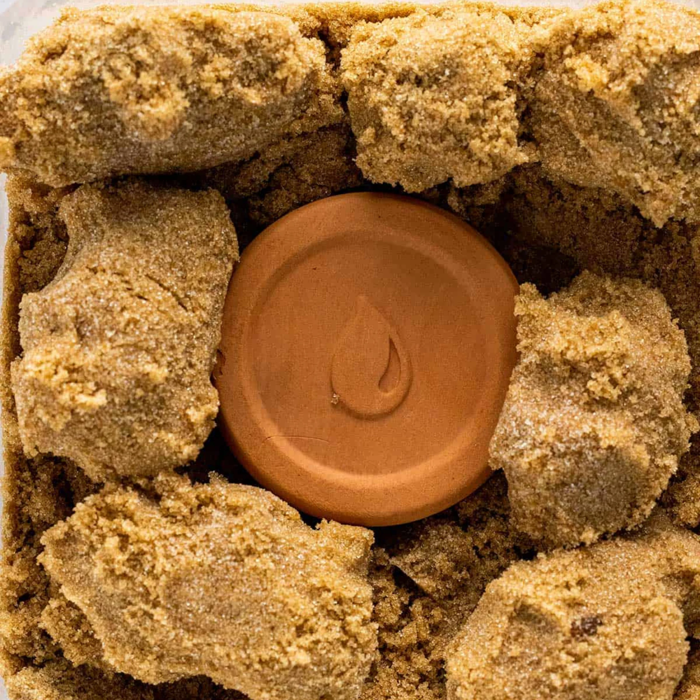 A terracotta-colored wax seal with an embossed droplet sits among rough, golden-brown chunks of natural material, creating a warm, earthy texture.