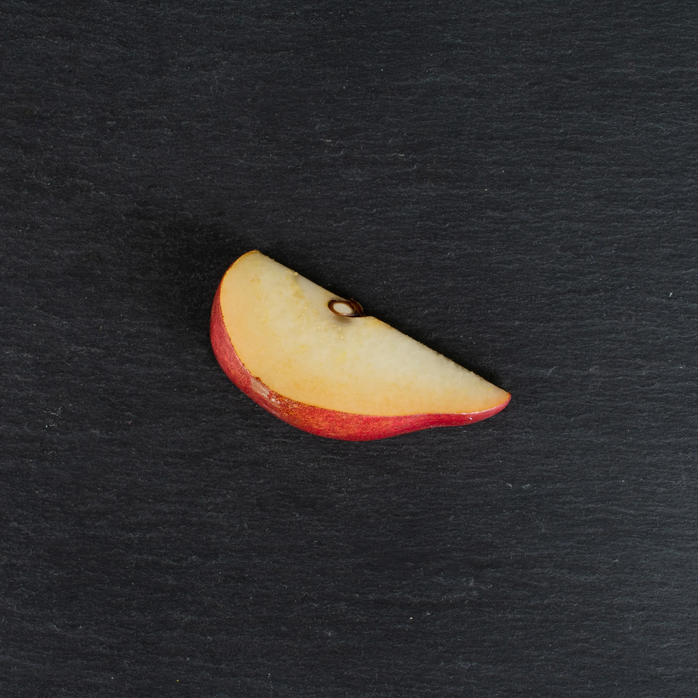 A single red-skinned apple slice with pale yellow flesh and a visible seed rests on a dark slate surface, creating a simple, quiet still life.
