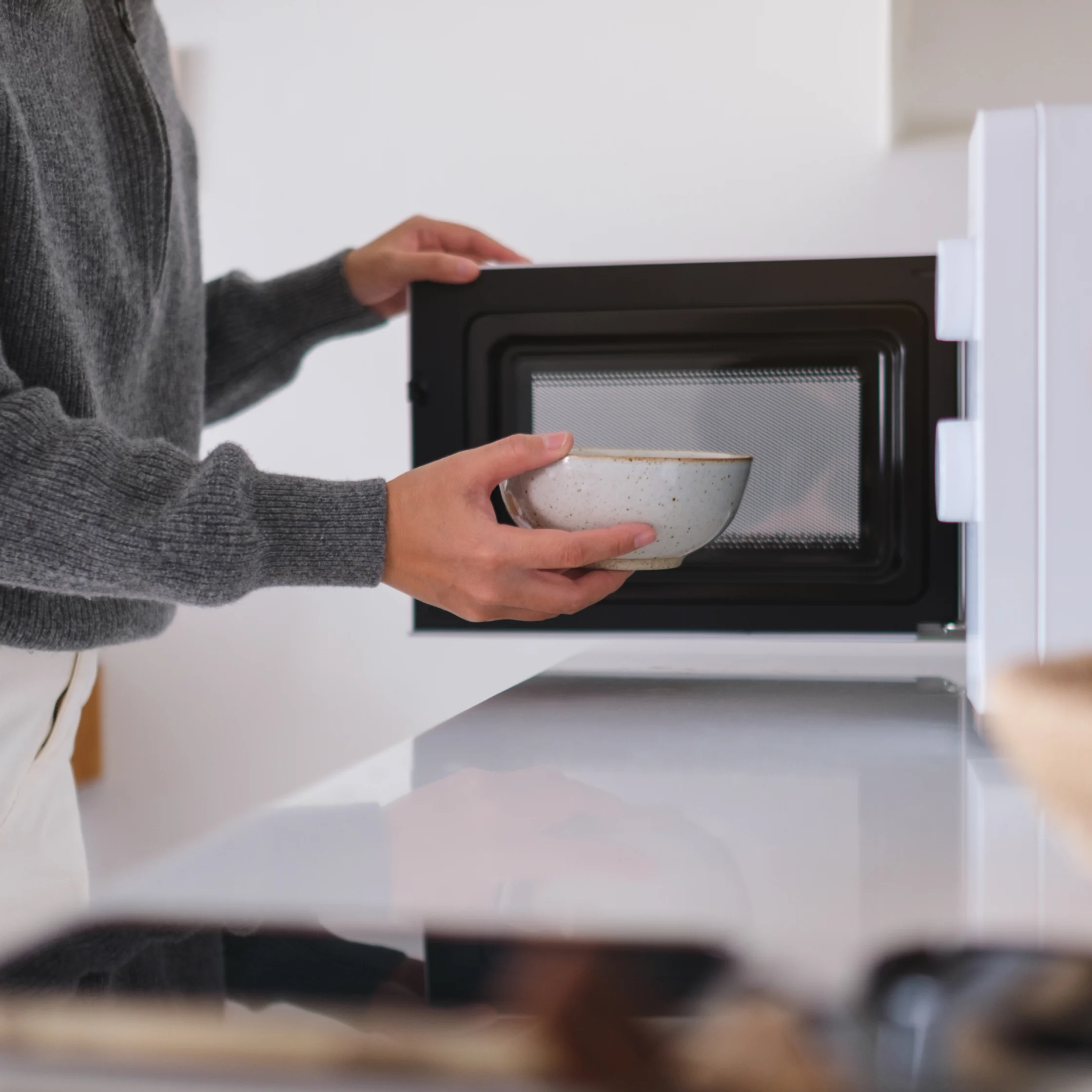 Person in a gray sweater places a white bowl into an open microwave in a bright kitchen, conveying a calm, everyday cooking moment.