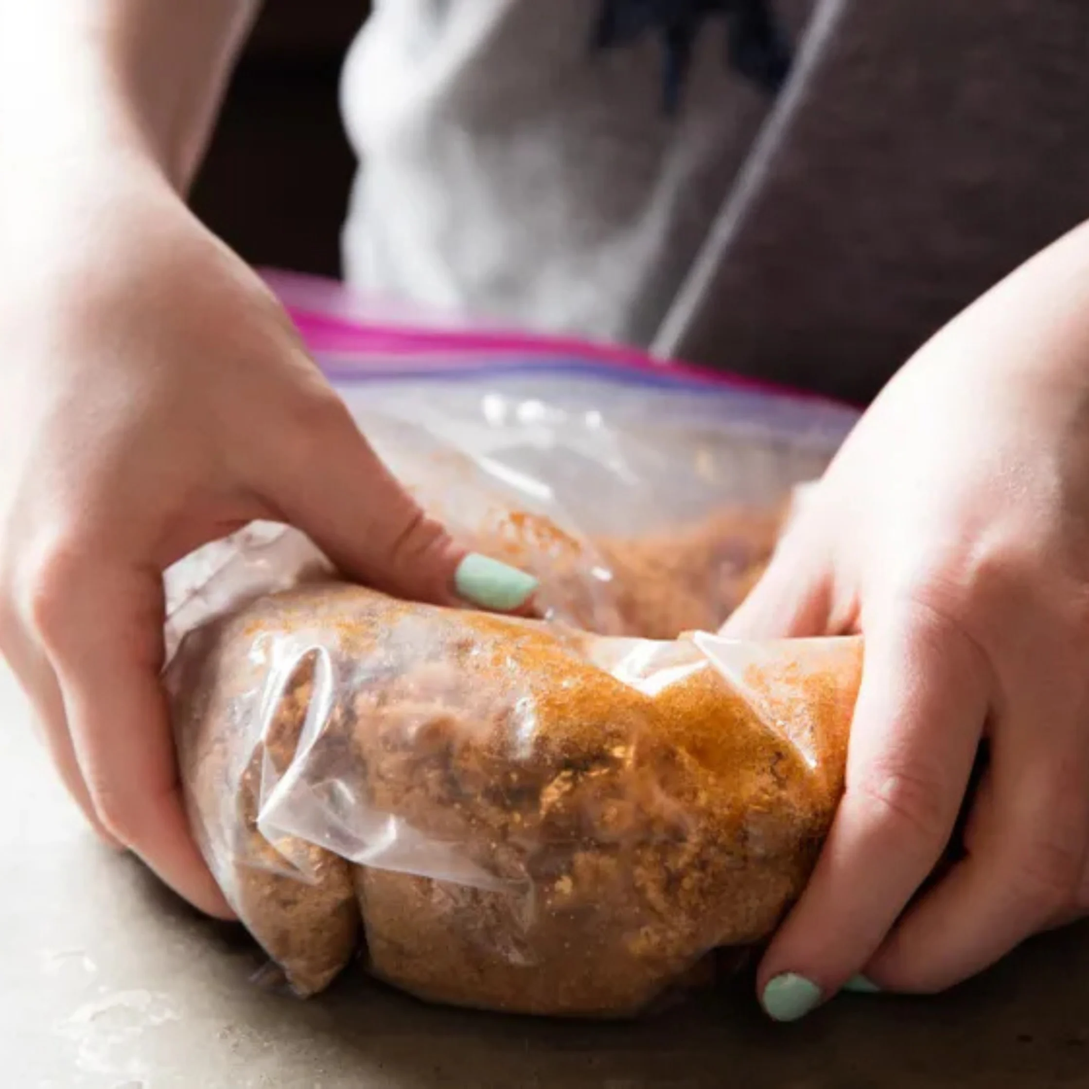 Hands with mint-green nail polish knead a seasoned mixture inside a clear zip-top bag, suggesting hands-on meal prep in a calm kitchen.