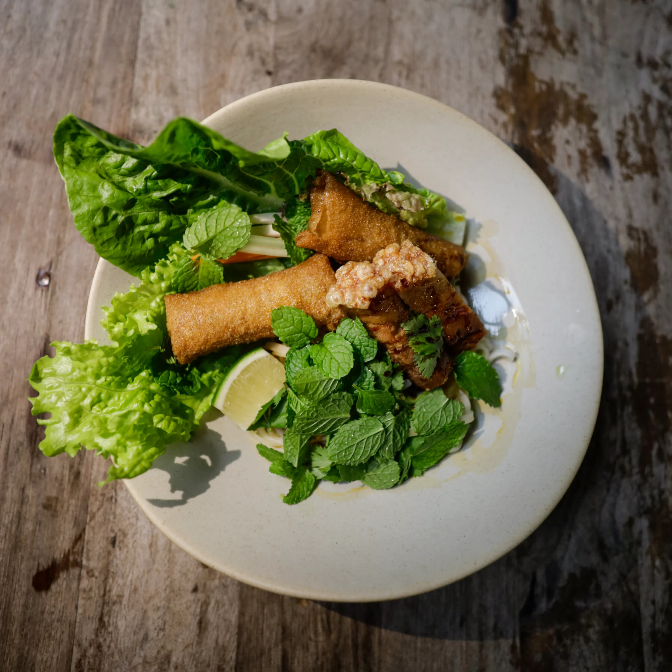 A plate of crispy golden spring rolls with browned bites, fresh lettuce, mint, and lime on a rustic wooden table, inviting and appetizing.