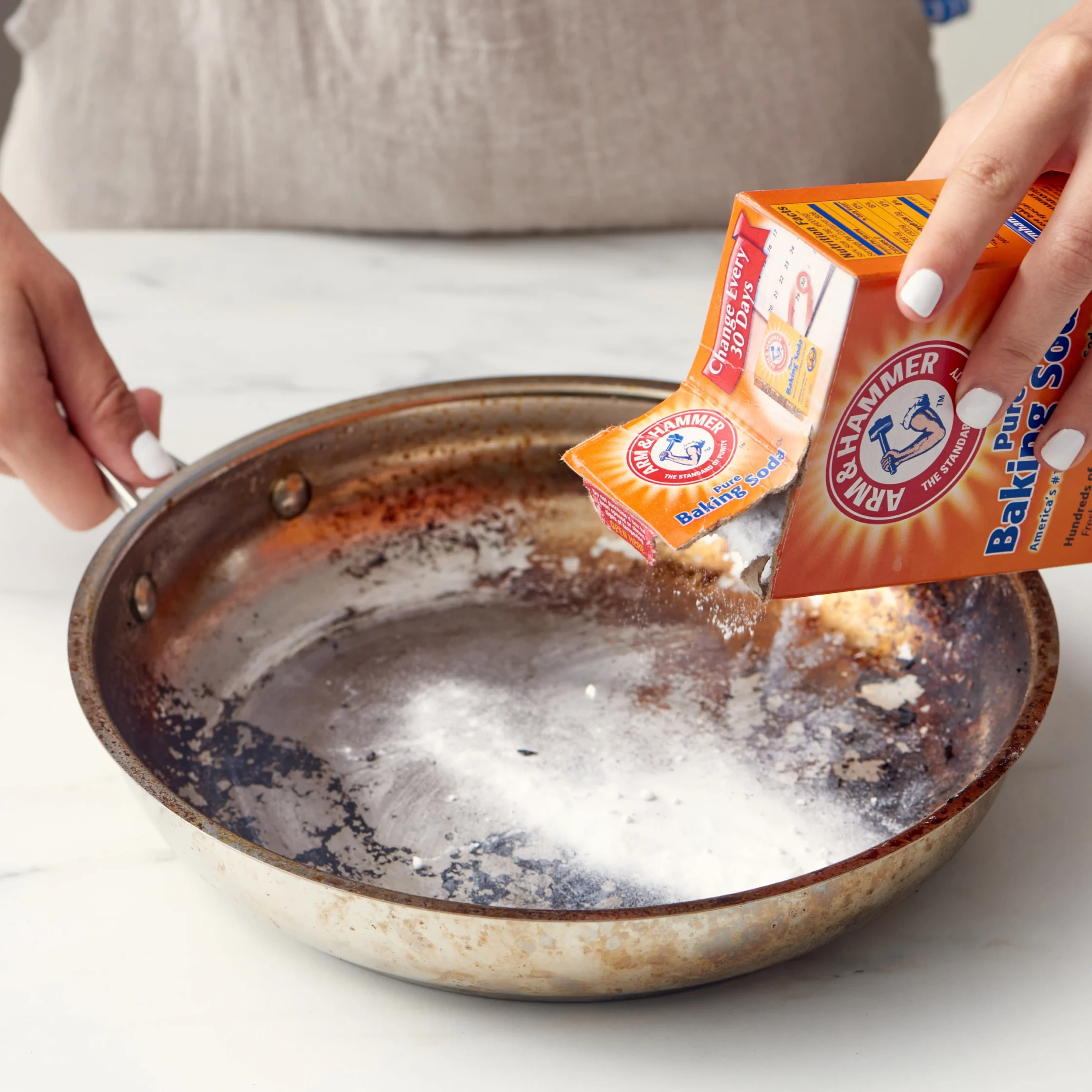 Hands pour baking soda from an Arm & Hammer box into a worn, greasy pan in a kitchen, suggesting a practical cleaning task.