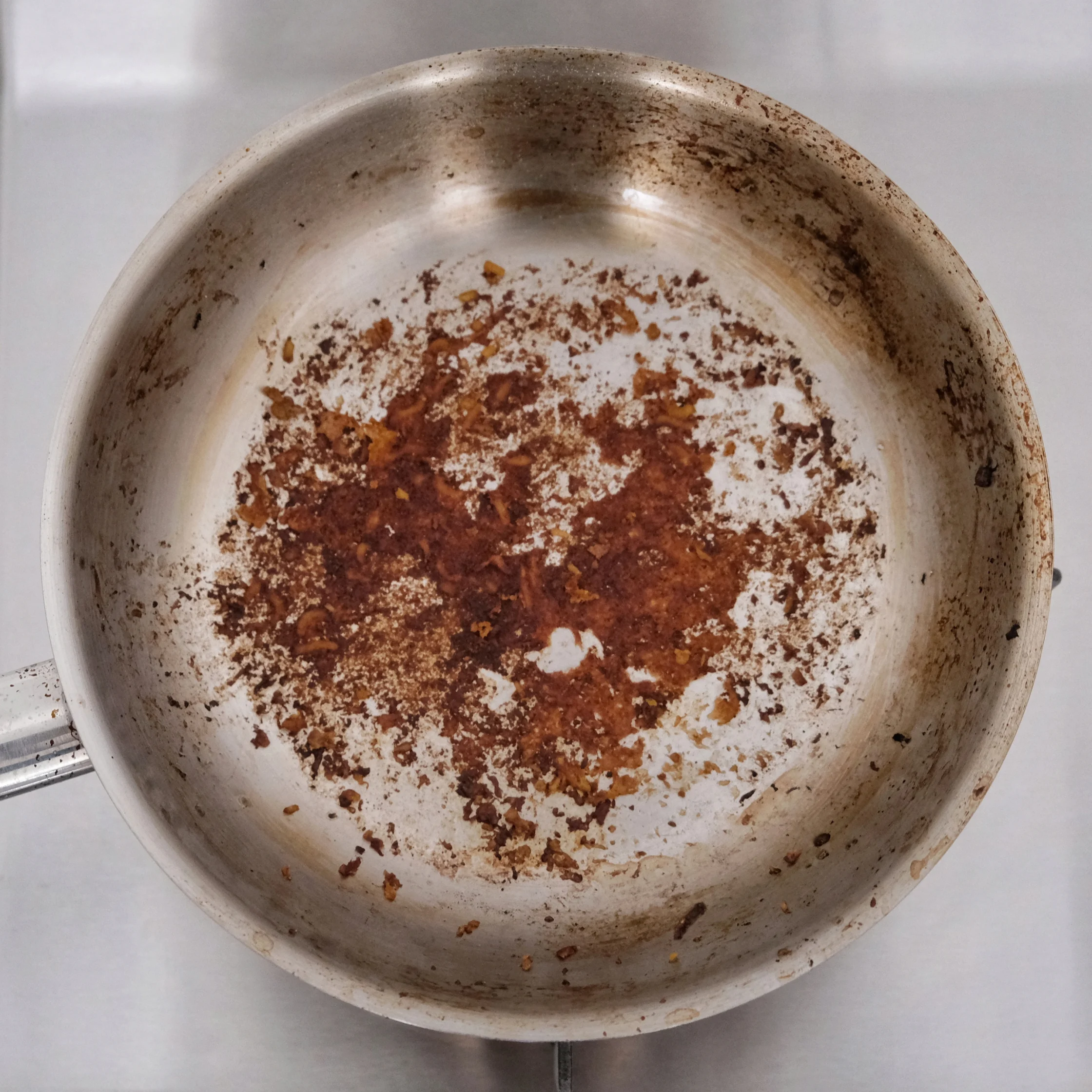 Top-down view of a well-used stainless-steel frying pan with dark, burnt-on brown food residue across its center and stained sides.