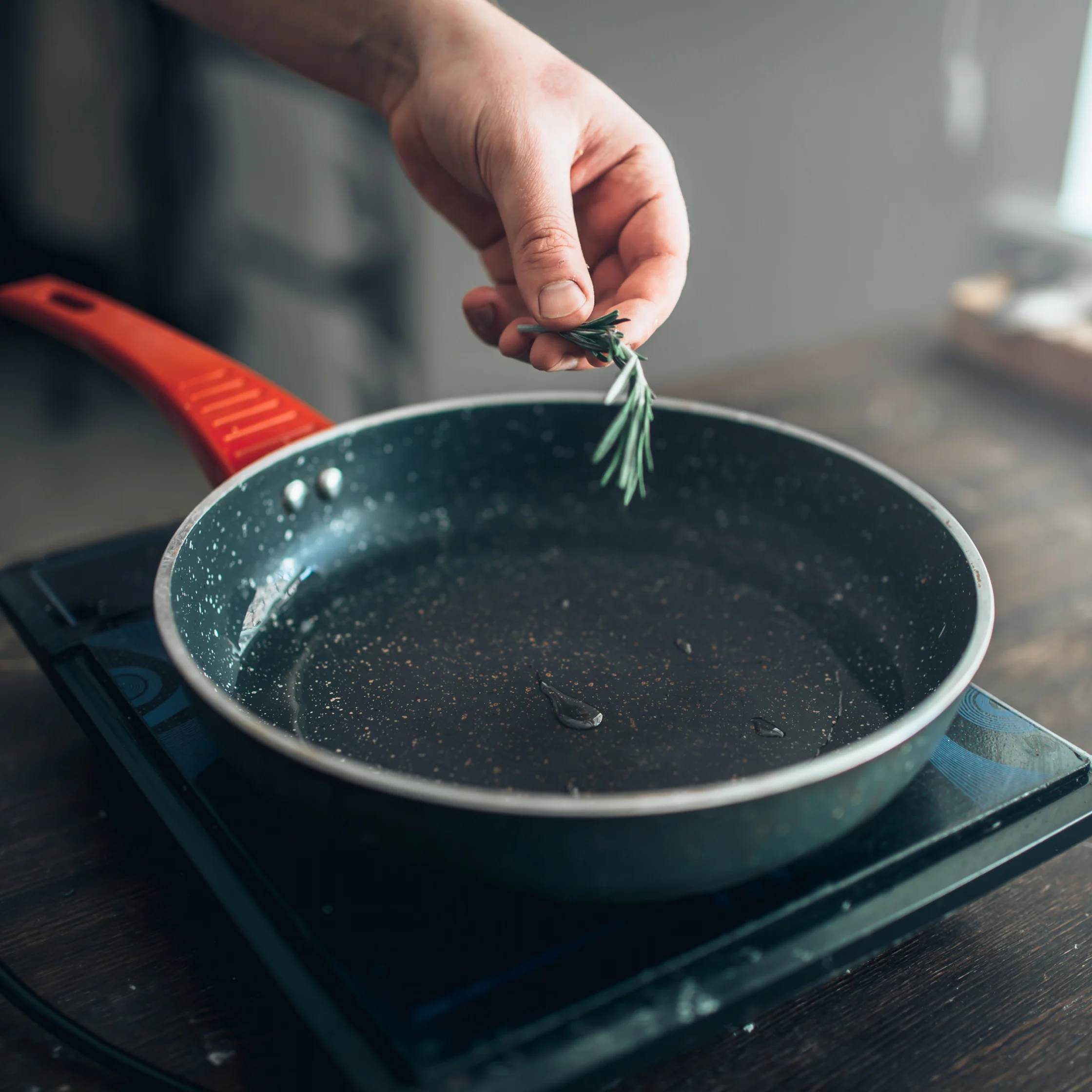 A hand sprinkles fresh rosemary into a dark frying pan on a stovetop, adding an aromatic touch to meal preparation in a calm kitchen.