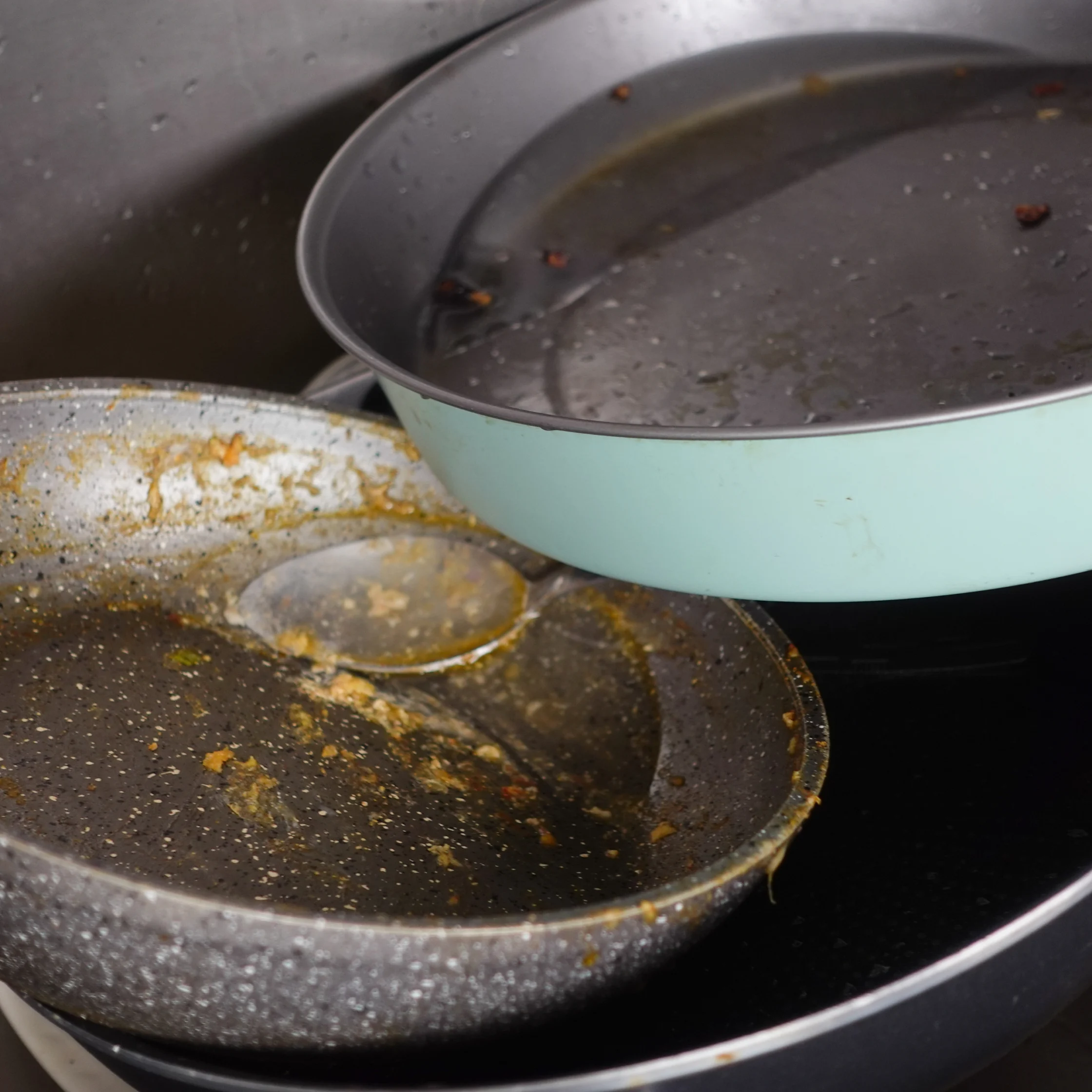 A dirty, speckled frying pan and pale blue pot sit stacked in a sink, with greasy residue and food scraps left behind, conveying a chore to tackle.