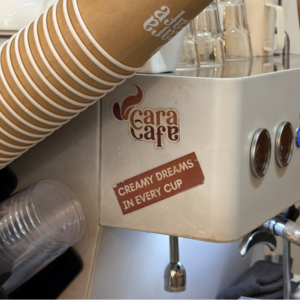 Close-up of a café beverage machine labeled “Cara Café” and “Creamy dreams in every cup,” with stacked paper cups and clear glasses, creating a warm, inviting feel.