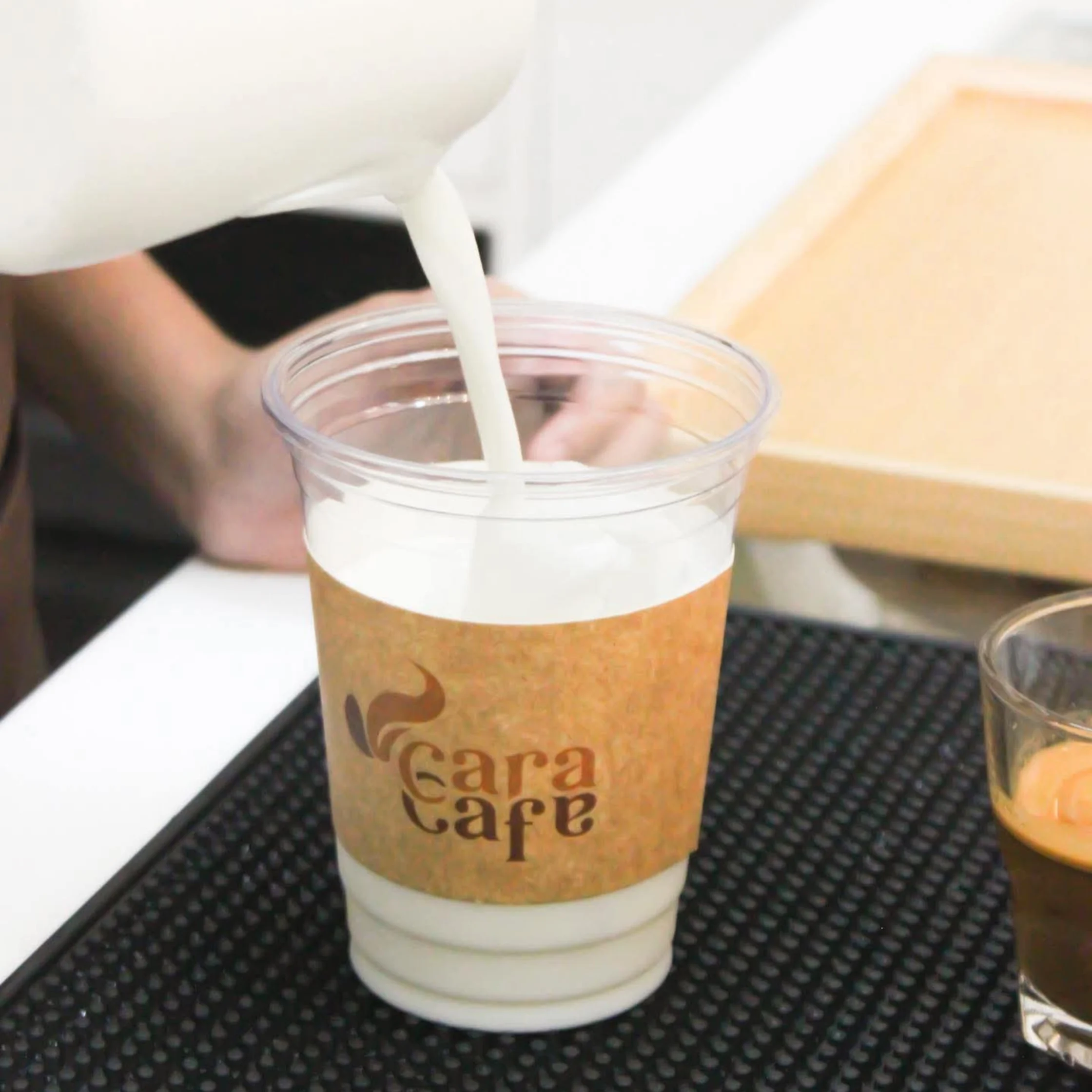 A barista pours milk into a clear plastic cup of coffee on a black counter mat; the Cara Cafe sleeve and warm, calm café setting are visible.