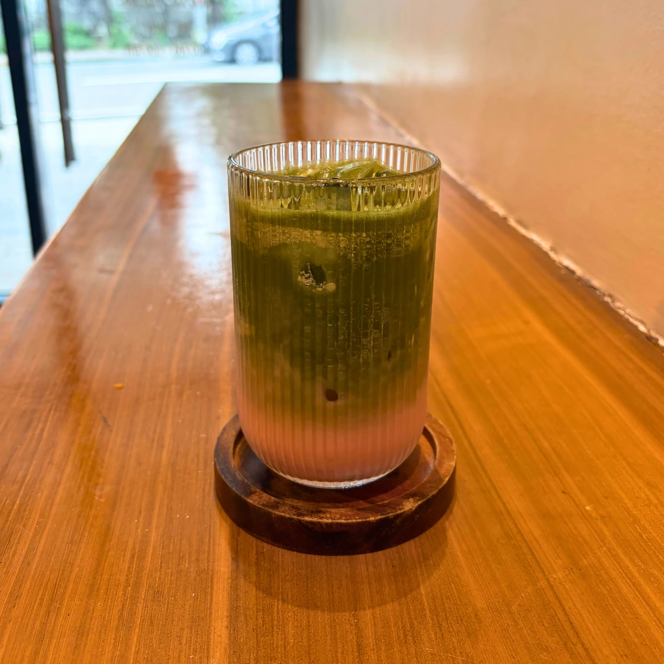 A layered iced green matcha drink in a ribbed glass sits on a wooden coaster atop a honey-colored café table, creating a calm, inviting scene.