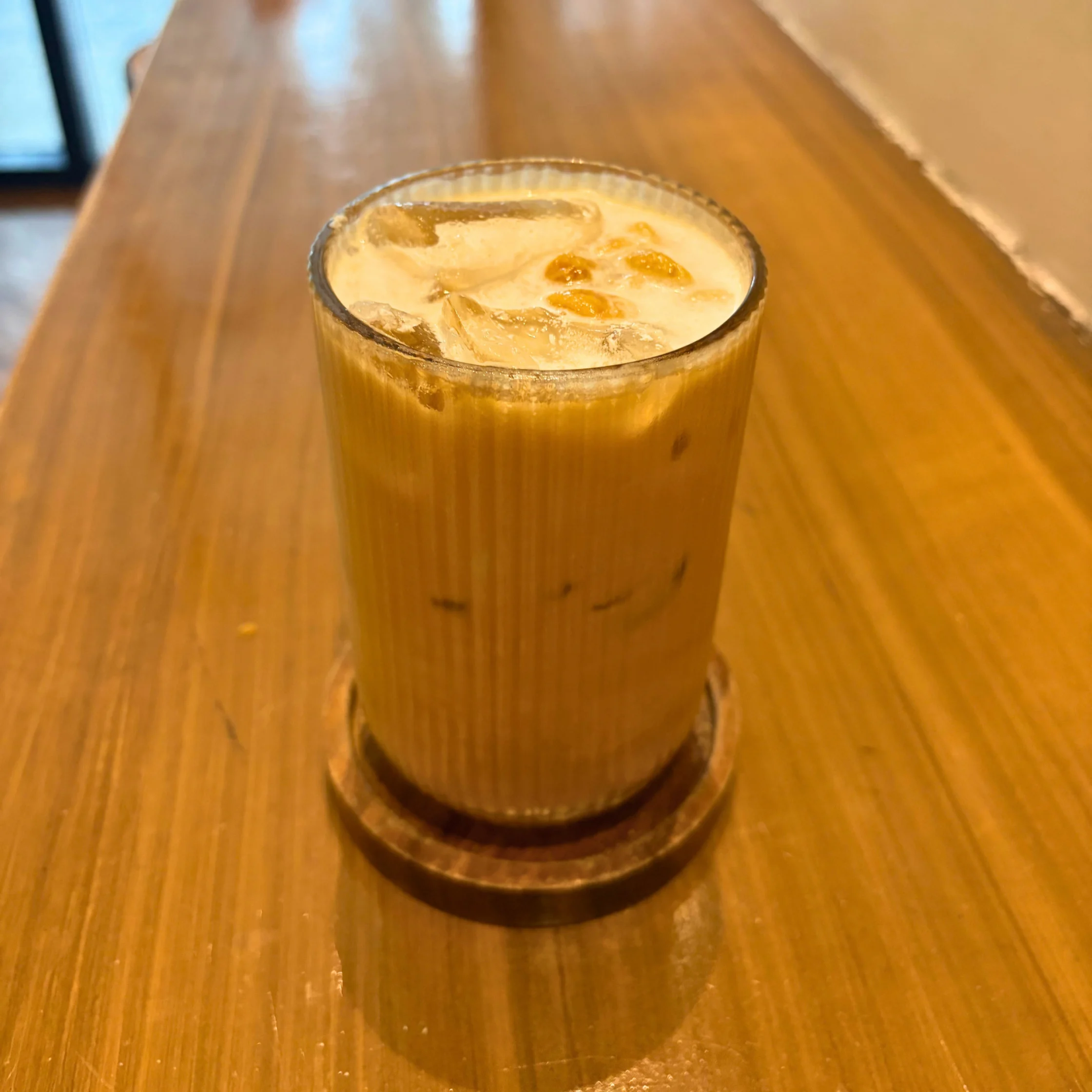 A chilled golden iced coffee topped with creamy foam and ice sits in a clear glass on a coaster atop a polished wooden table, inviting a relaxed café moment.
