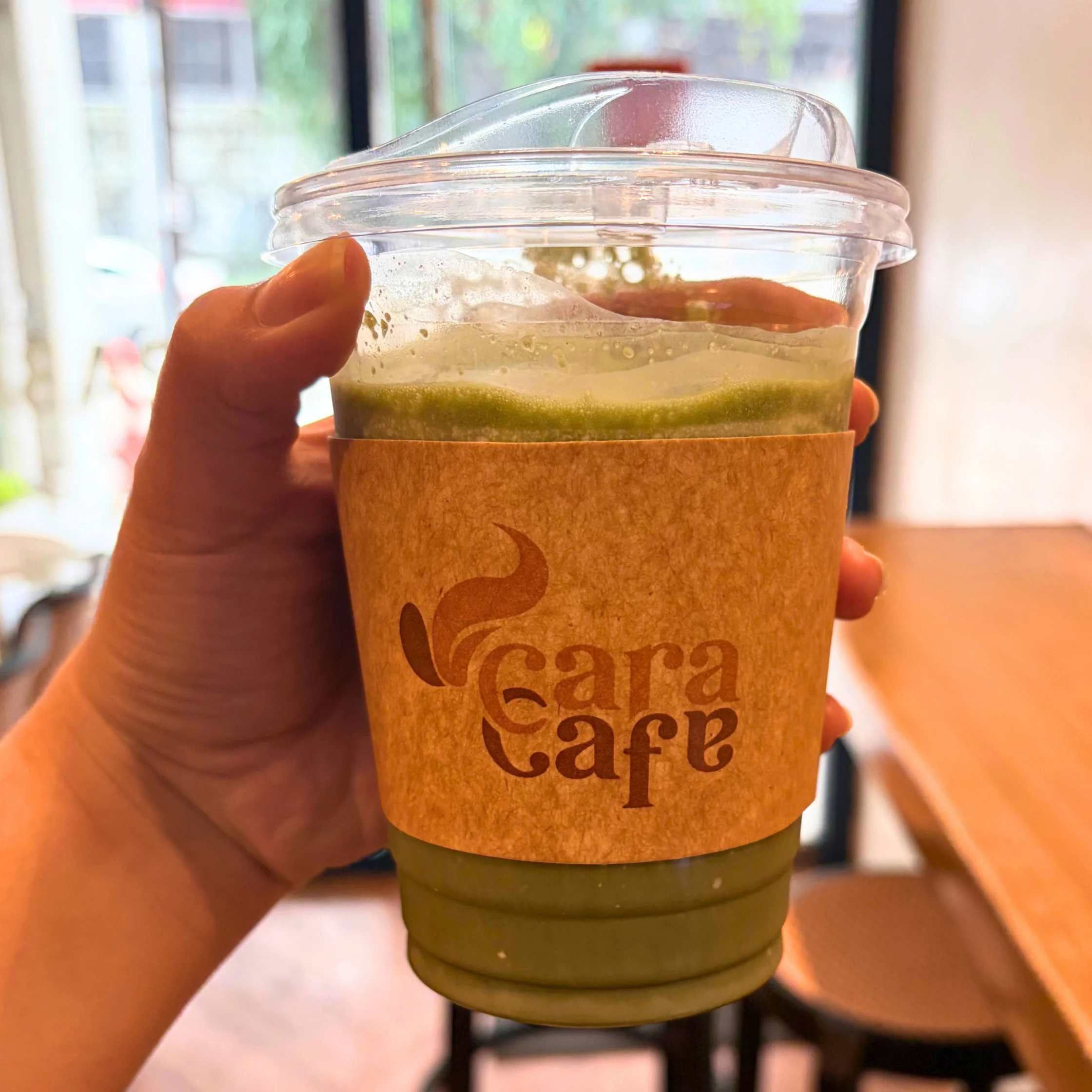 A hand holds a clear takeaway cup of layered green matcha with a brown cardboard sleeve marked “Cara Café,” in a bright café; warm, inviting mood.