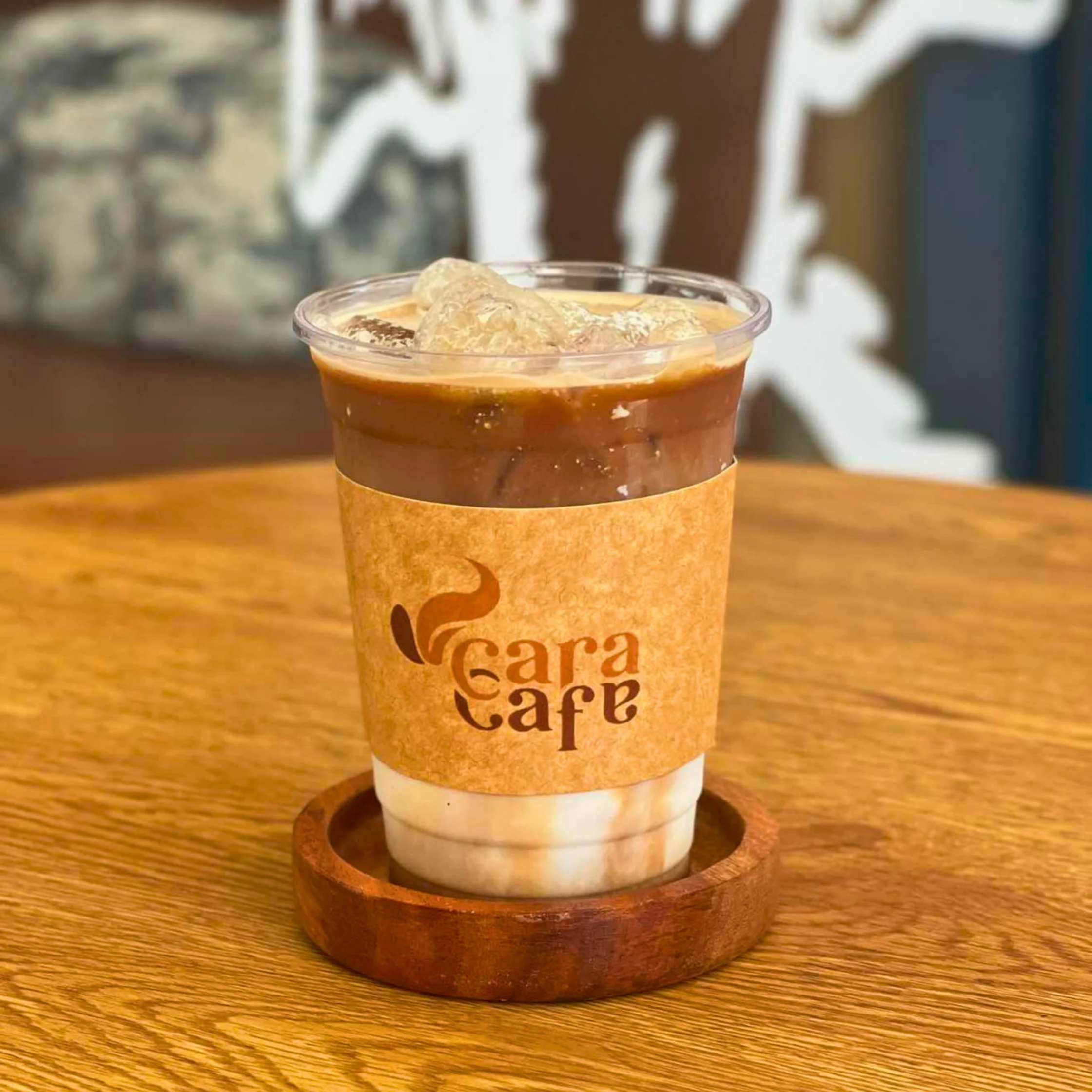 An iced brown coffee drink with layered milk sits in a clear Cara Café cup on a wooden coaster and table, creating a warm, inviting café scene.