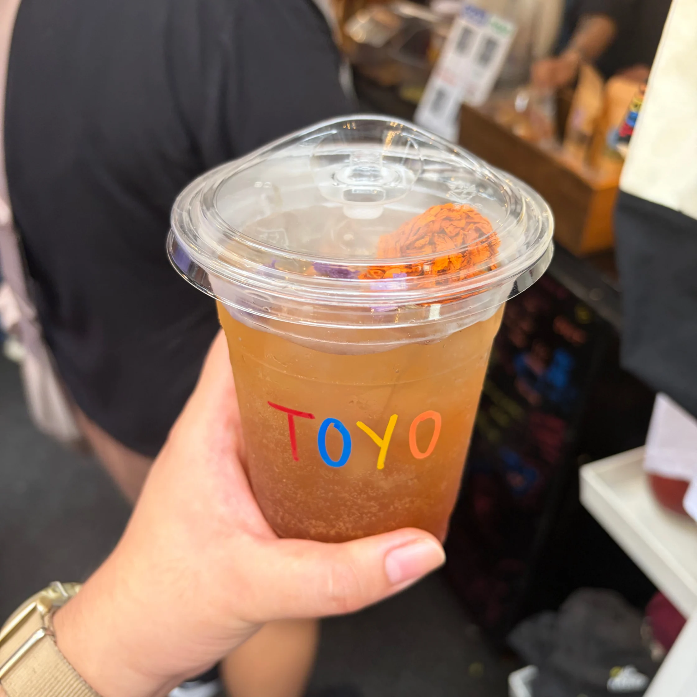 A hand holds a clear plastic cup of amber bubble tea with colorful “TOYO” lettering, a domed lid, and orange topping, amid a busy shop counter.