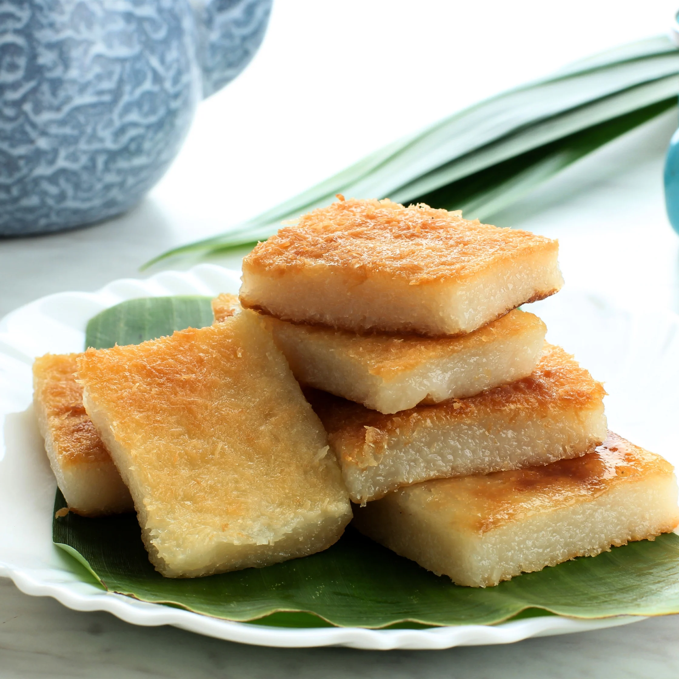 Golden-brown grilled cassava cakes are stacked on a banana leaf-lined white plate, with a patterned blue bowl and green leaves behind them; the scene feels warm and inviting.