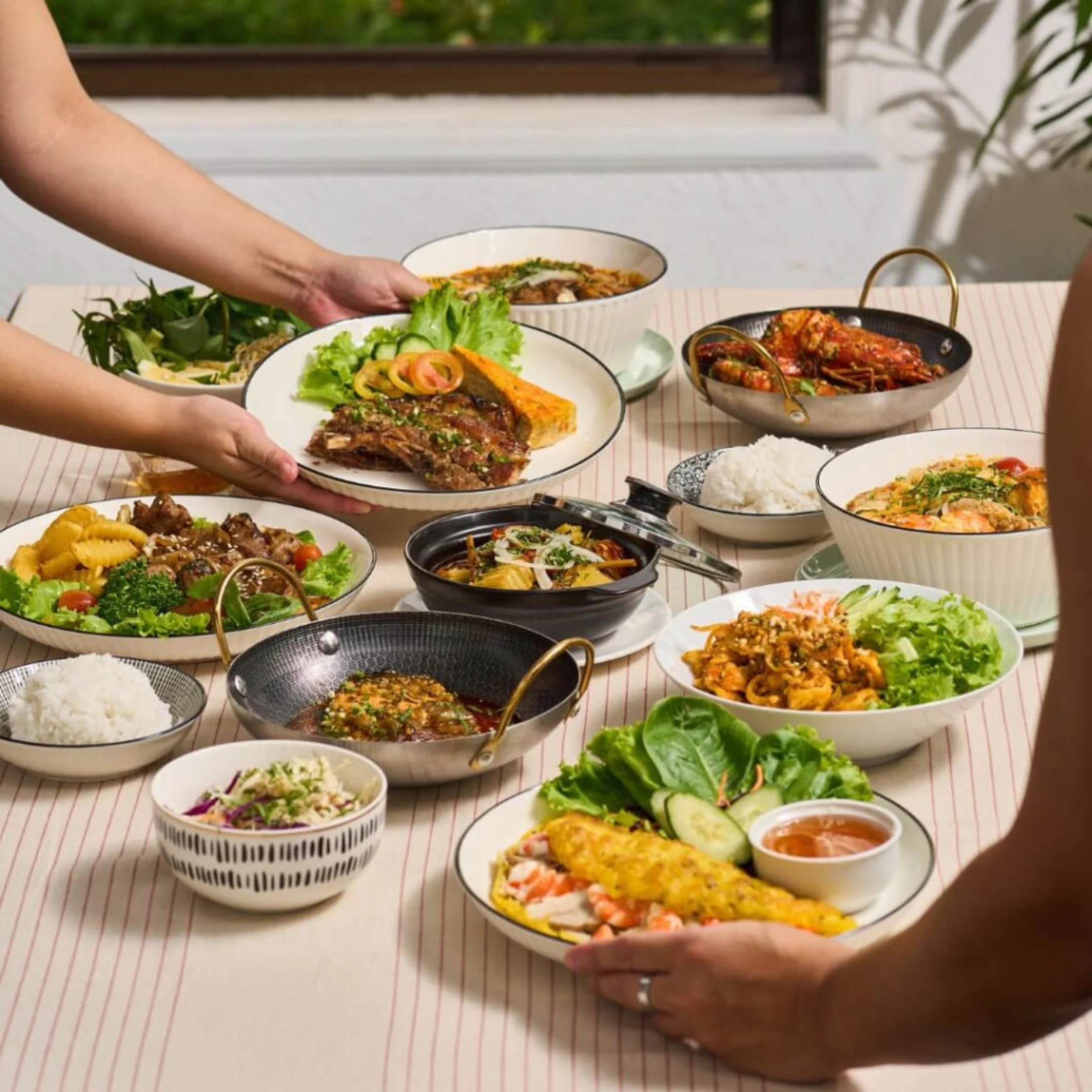 Hands serve a colorful Asian-style meal on a striped outdoor table, with rice, grilled fish, stir-fried dishes, salads, and dipping sauces.