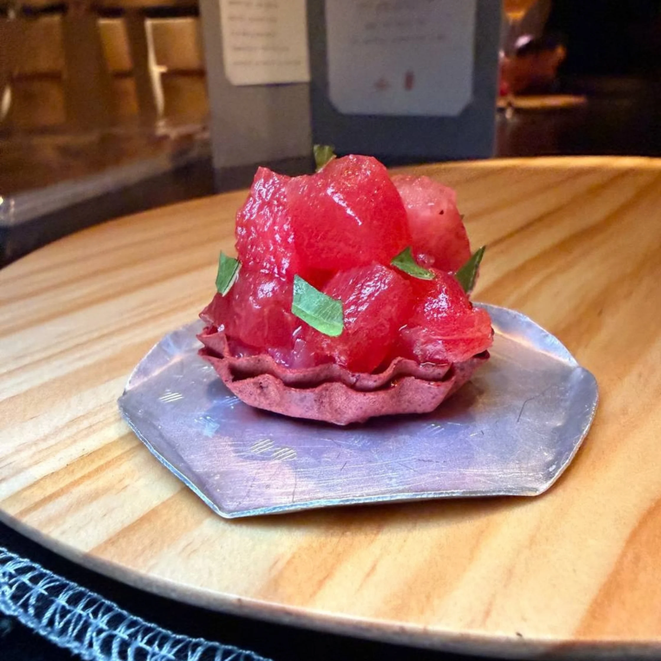 A glossy red fruit tart topped with fresh fruit and green leaves sits on a metal plate atop a wooden counter, creating an inviting dessert display.