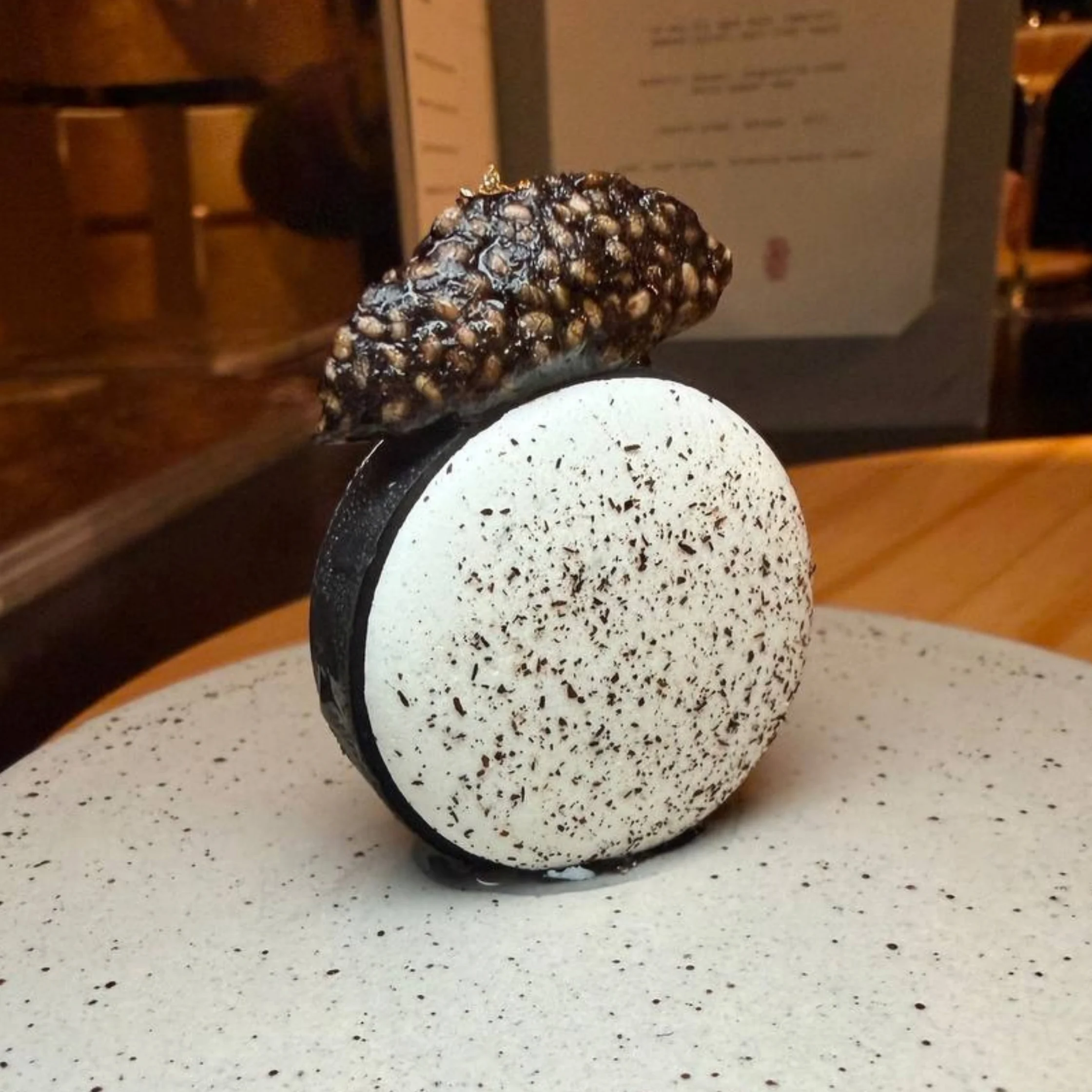 A sculptural dessert with a white, black-speckled round base and glossy, textured dark topping sits on a ceramic plate in a warmly lit restaurant.