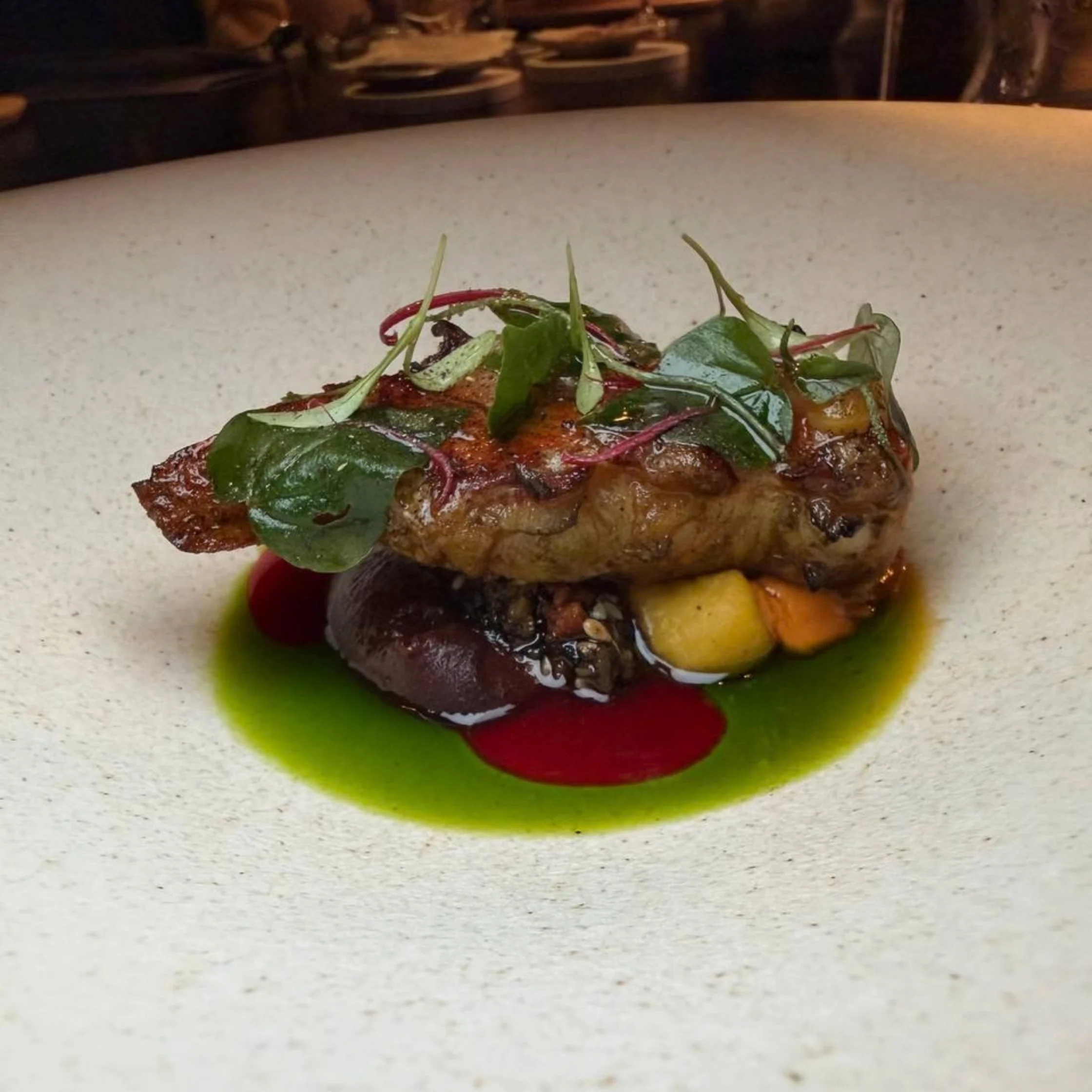 A seared meat entrée topped with fresh greens, served over vivid green herb sauce with red and dark purées and golden vegetables on a white plate.